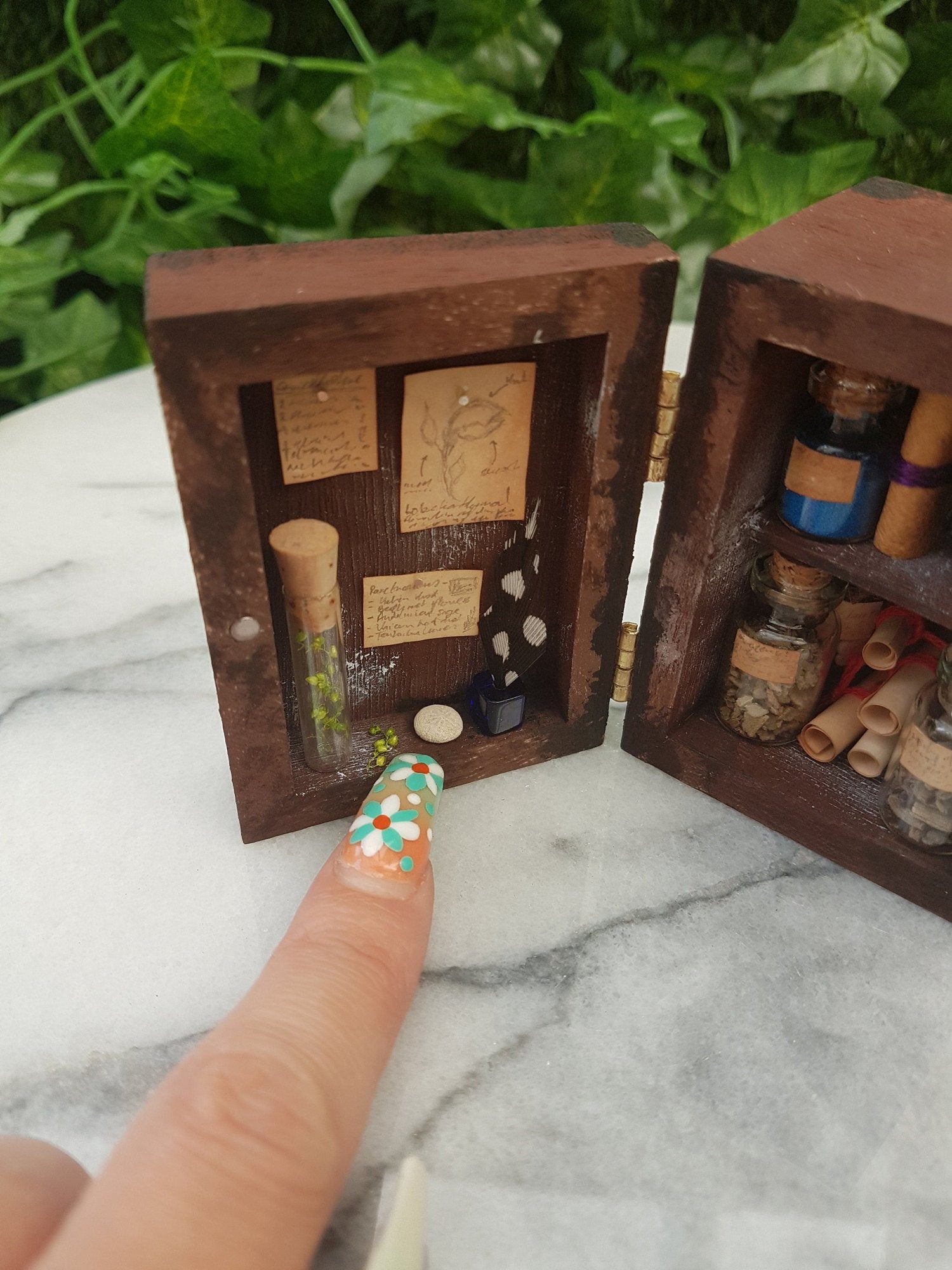 1:12 scale miniature brown apothecary cabinet is shown open. Shelves are filled with glass vials, parchment scrolls, cauldron and leather book. The left cabinet door has aged notes pinned up with an inkwell and quill. A finger points for scale