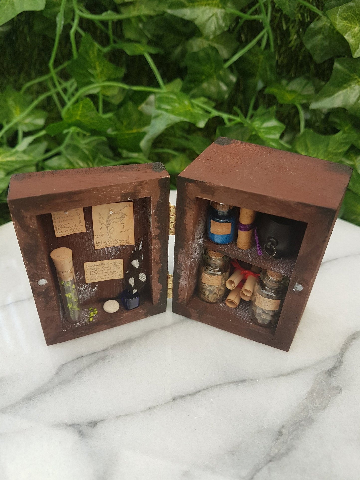 1:12 scale miniature brown apothecary cabinet is shown open on a marble slab with shelves filled with glass vials, parchment scrolls, miniature cauldron and leather bound book. The left cabinet door has aged notes pinned up with an inkwell and quill