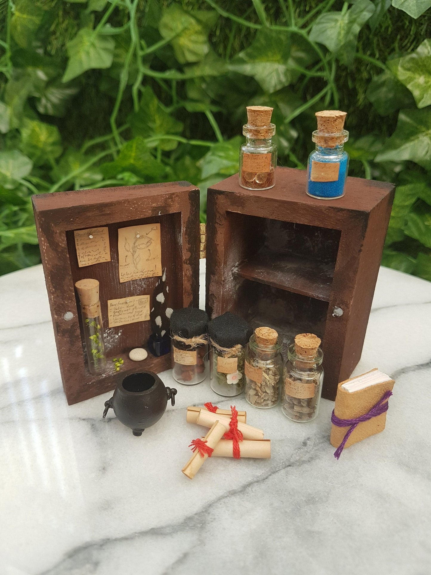 A 1:12 scale miniature brown apothecary cabinet is shown open on a marble slab with glass vials, parchment scrolls, miniature cauldron and leather bound book. The left cabinet door has aged notes pinned up with an inkwell and quill