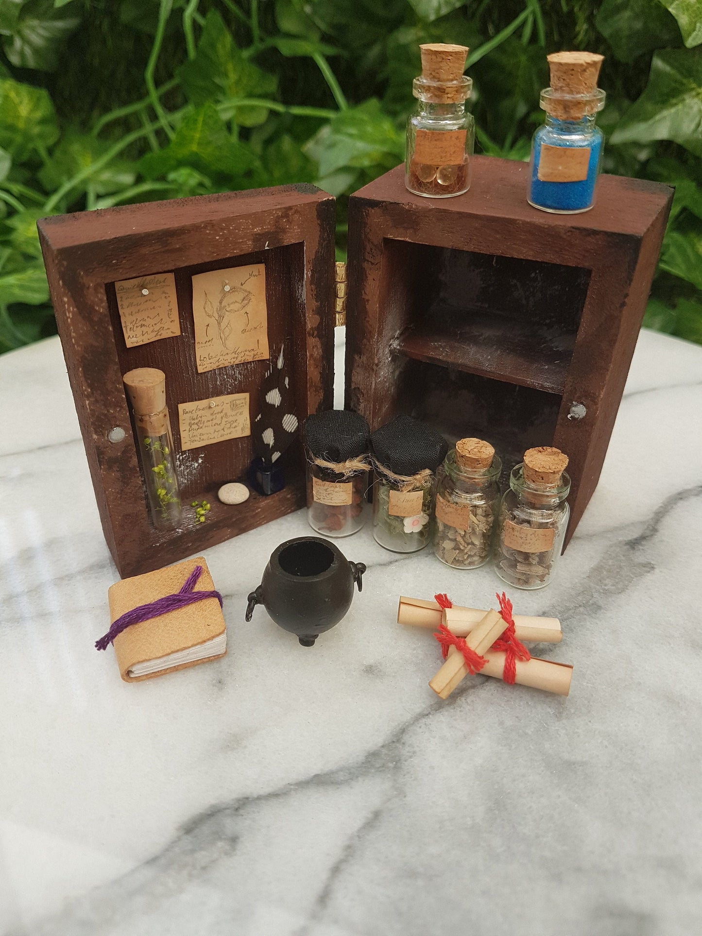 A 1:12 scale miniature brown apothecary cabinet is shown open on a marble slab with glass vials, parchment scrolls, miniature cauldron and leather bound book. The left cabinet door has aged notes pinned up with an inkwell and quill