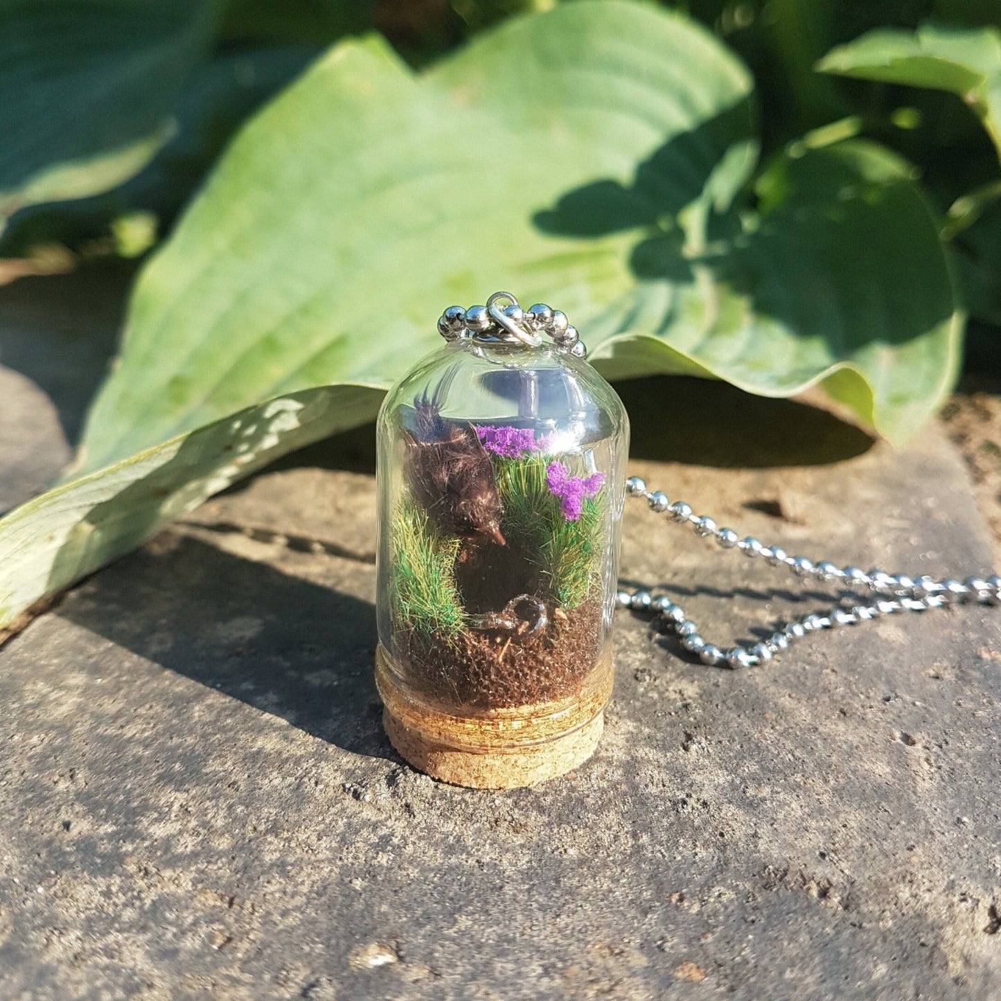 Miniature diorama glass bell jar necklace with cork base contains a tiny brown Robin. The Robin is surrounded by grass and purple flowers looking into an earthy hole containing a rusty key. The necklace is displayed on a grey stone with green leaves