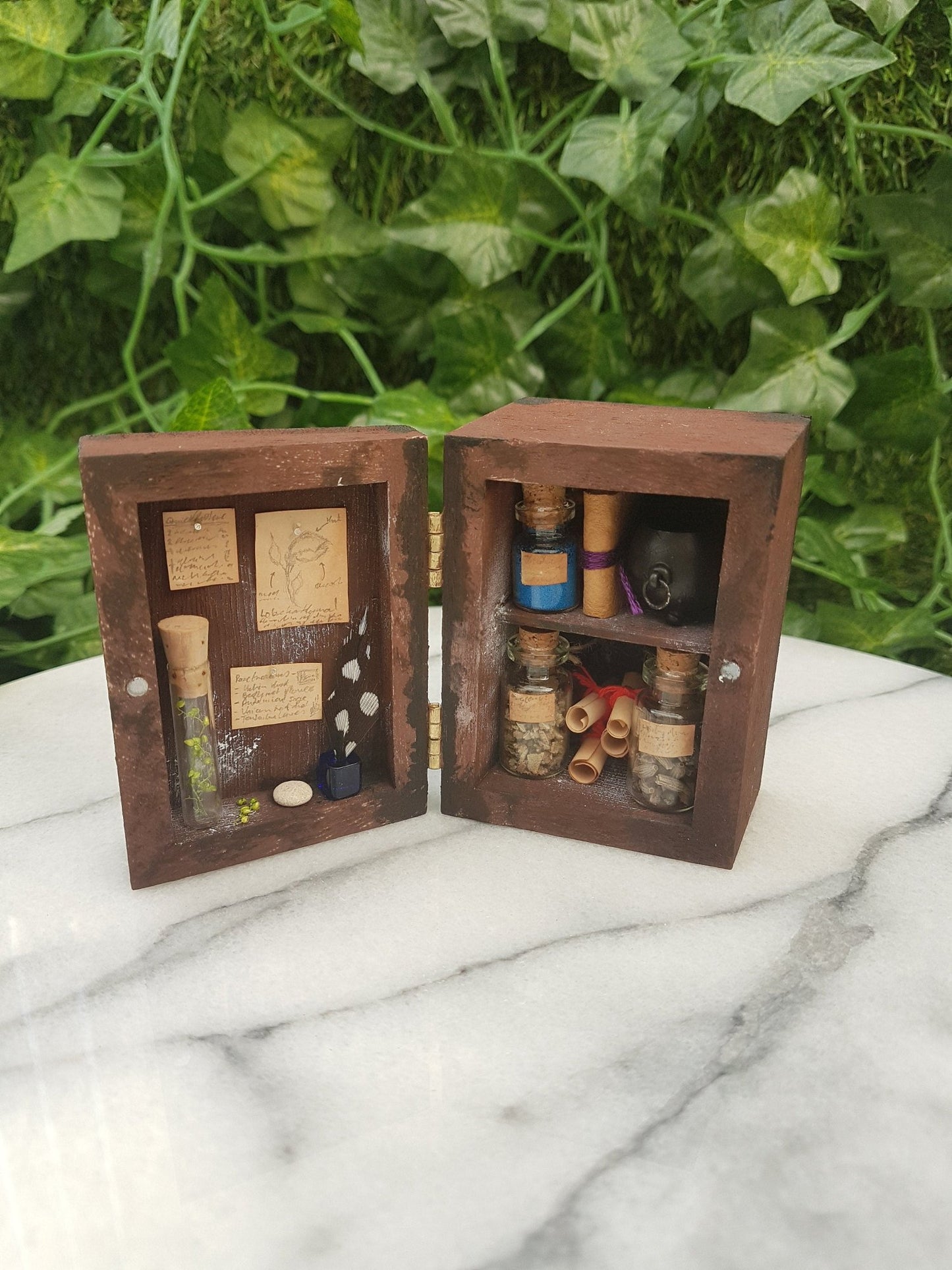 1:12 scale miniature brown apothecary cabinet is shown open on a marble slab with shelves filled with glass vials, parchment scrolls, miniature cauldron and leather bound book. The left cabinet door has aged notes pinned up with an inkwell and quill