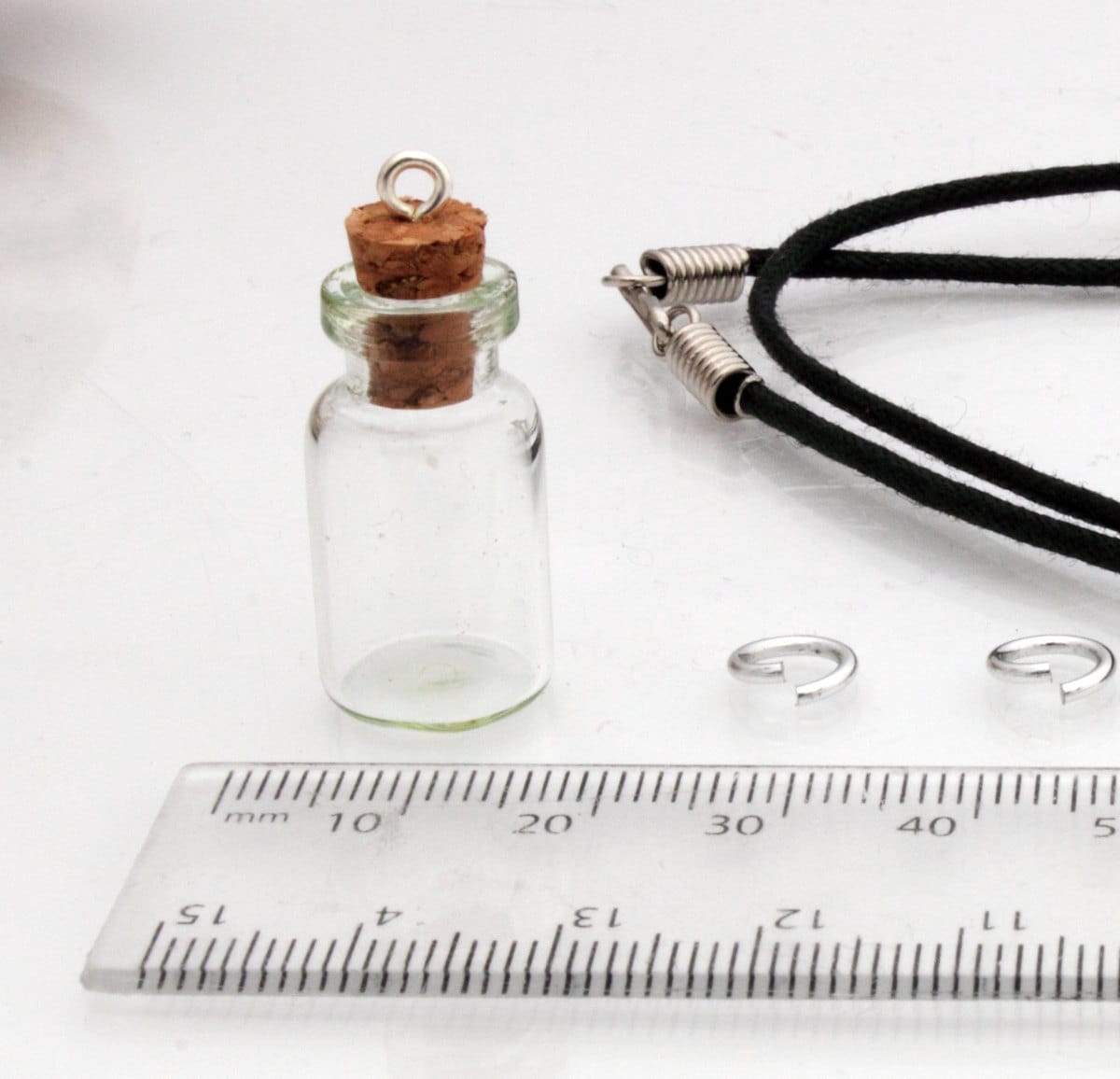 Medium jar glass vial with a cork bung sits beside a ruler, jump rings and a black pendant cord 