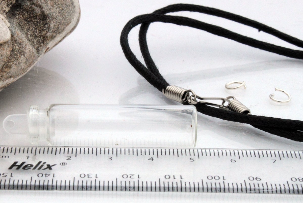 Tall glass vial with a plastic lid sits beside a ruler, jump rings and a black pendant cord 