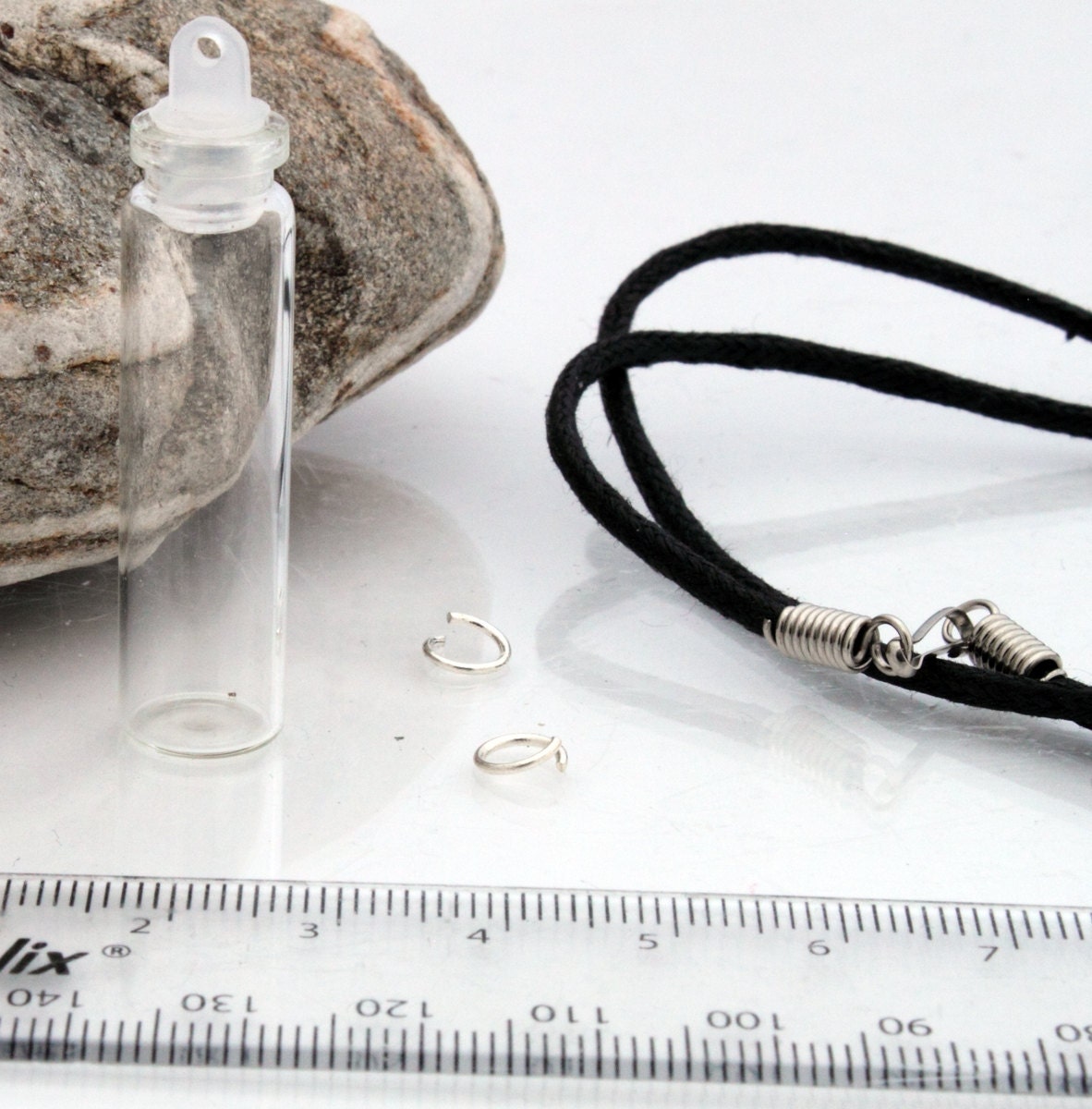 Tall glass vial with a plastic lid sits beside a ruler, jump rings and a black pendant cord 