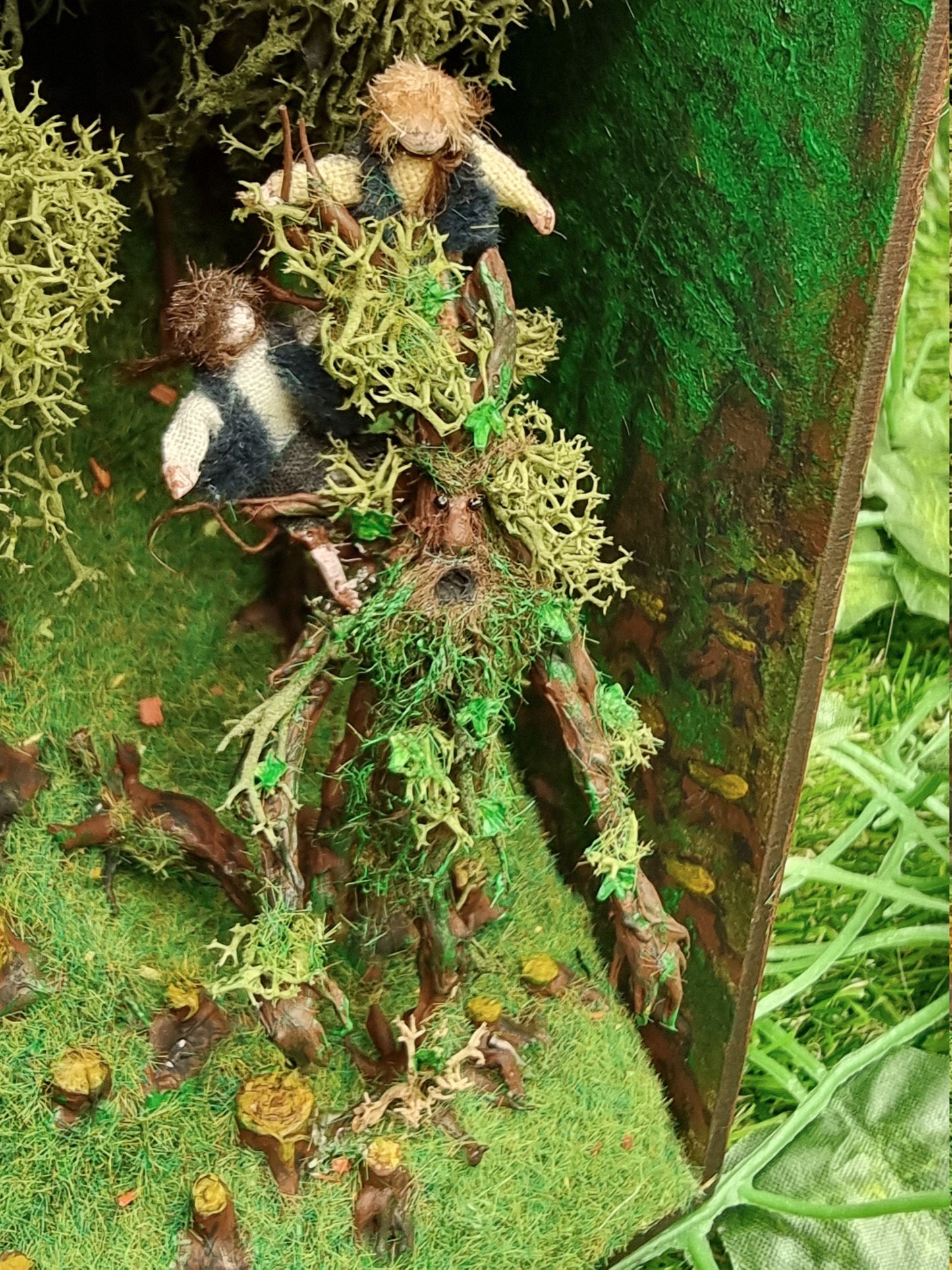 Miniature hobbits are seen sat on a living tree, Treebeard, in the booknook scene. The hobbits have green waistcoats with white, black and brown clothing. Treebeard has a green foliage beard and other organic parts. Treebeard has an open mouth