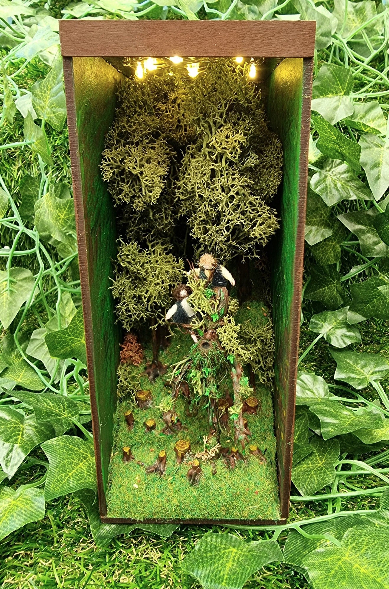 A small open fronted book nook shelf insert box containing fairy lights in the roof, a green grassy hill with thick tall trees at the back and tree stumps at the front. 2 hobbits are seen atop a living tree, treebeard. Green ivy background