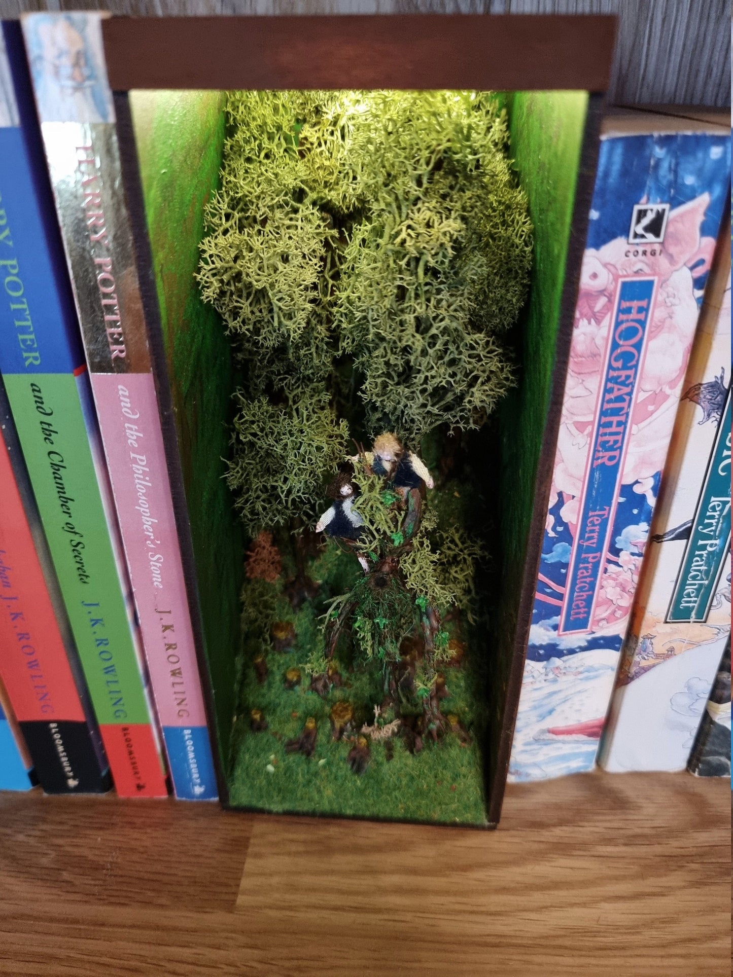 A small open fronted book nook shelf insert box sits between books of the same size on a bookshelf. Scene shows a green grassy hill with thick tall trees at the back and tree stumps at the front. 2 hobbits are seen atop a living tree, treebeard.