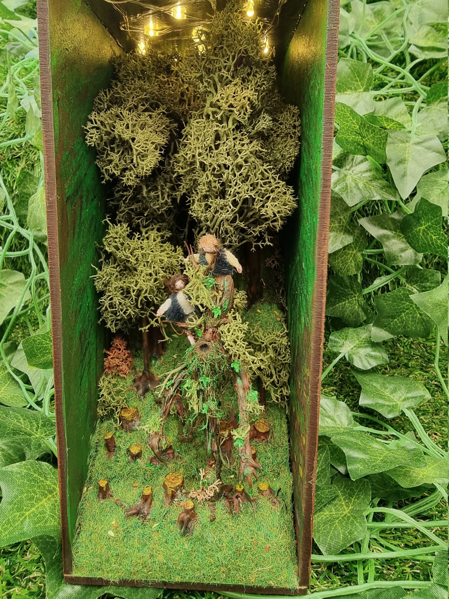 A small open fronted book nook shelf insert box containing fairy lights in the roof, a green grassy hill with thick tall trees at the back and tree stumps at the front. 2 hobbits are seen atop a living tree, treebeard. Green ivy background