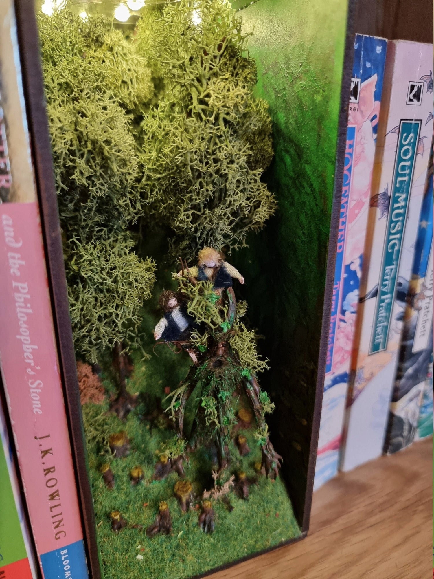 A small open fronted book nook shelf insert box sits between books of the same size on a bookshelf. The box contains a green grassy hill with thick tall trees at the back and tree stumps at the front. 2 hobbits are seen atop a living tree, treebeard.