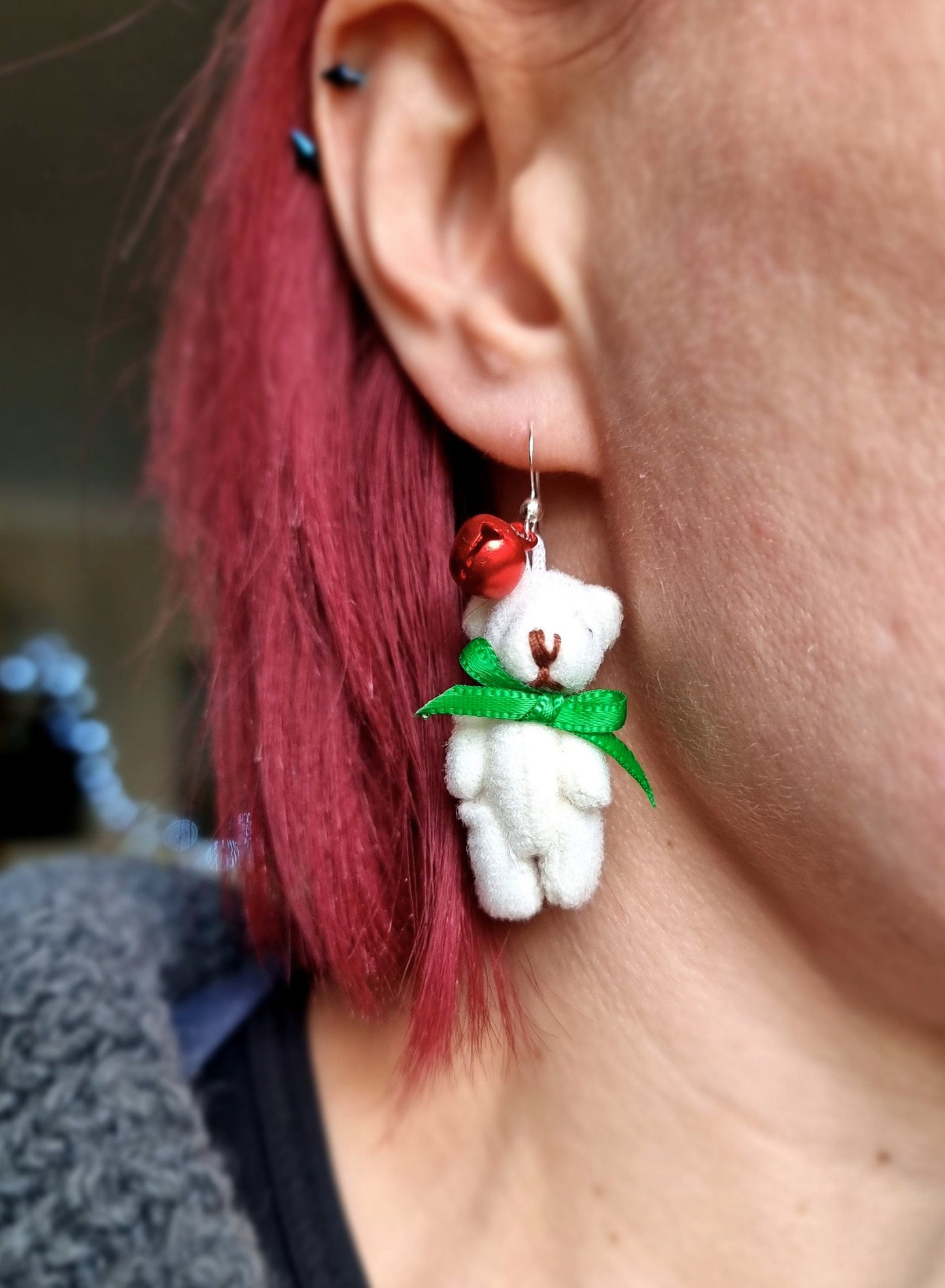 Miniature cream teddy bear earring with red bell and green bow tie hand from the ear lobe of a person with purple hair