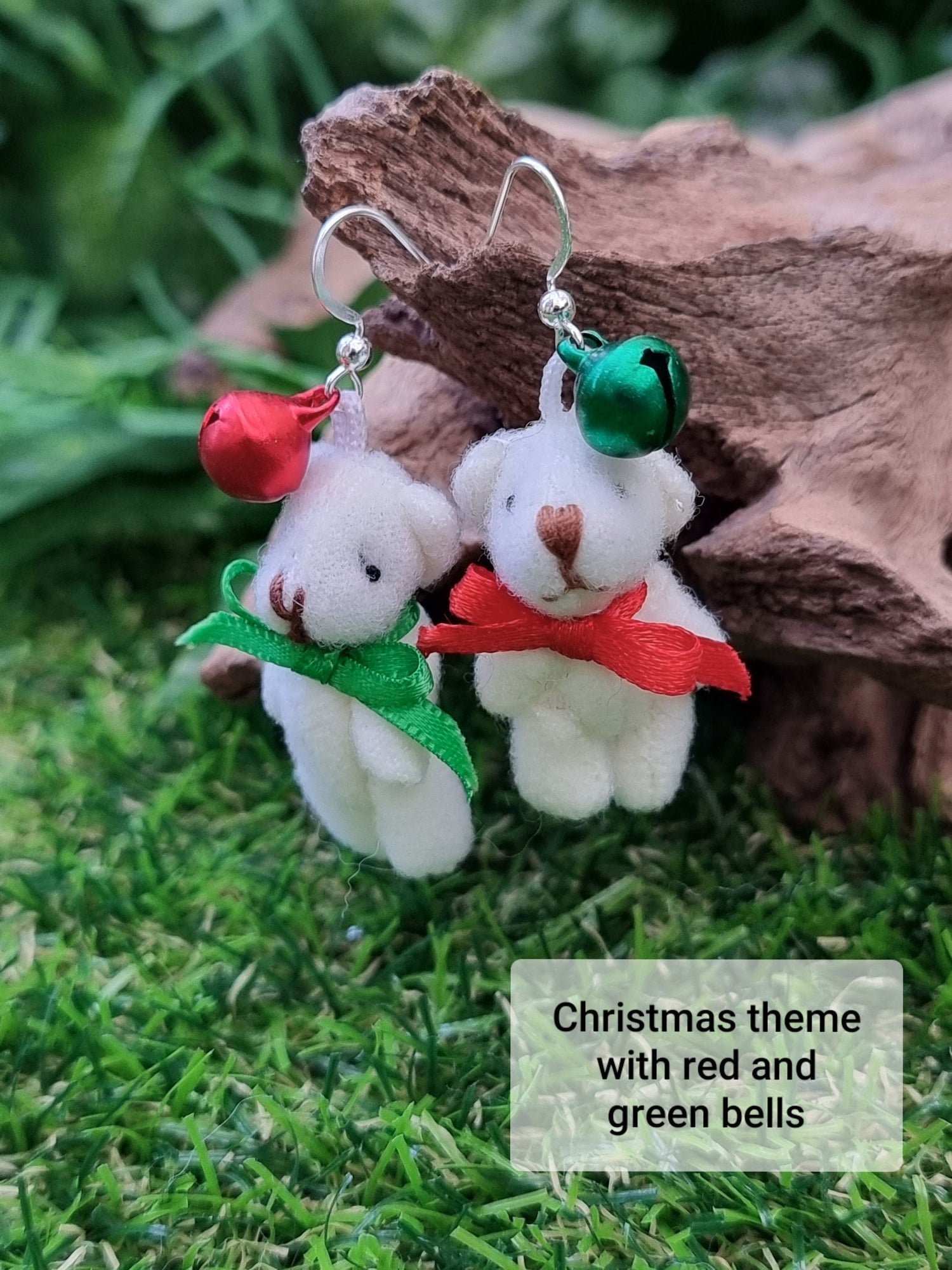 Two silver coloured hook style earrings hang from driftwood. One hook has a small green bell and one has a small red bell. Each hook has a cream coloured fabric teddy bear with an alternate coloured ribbon bow tie to the bell