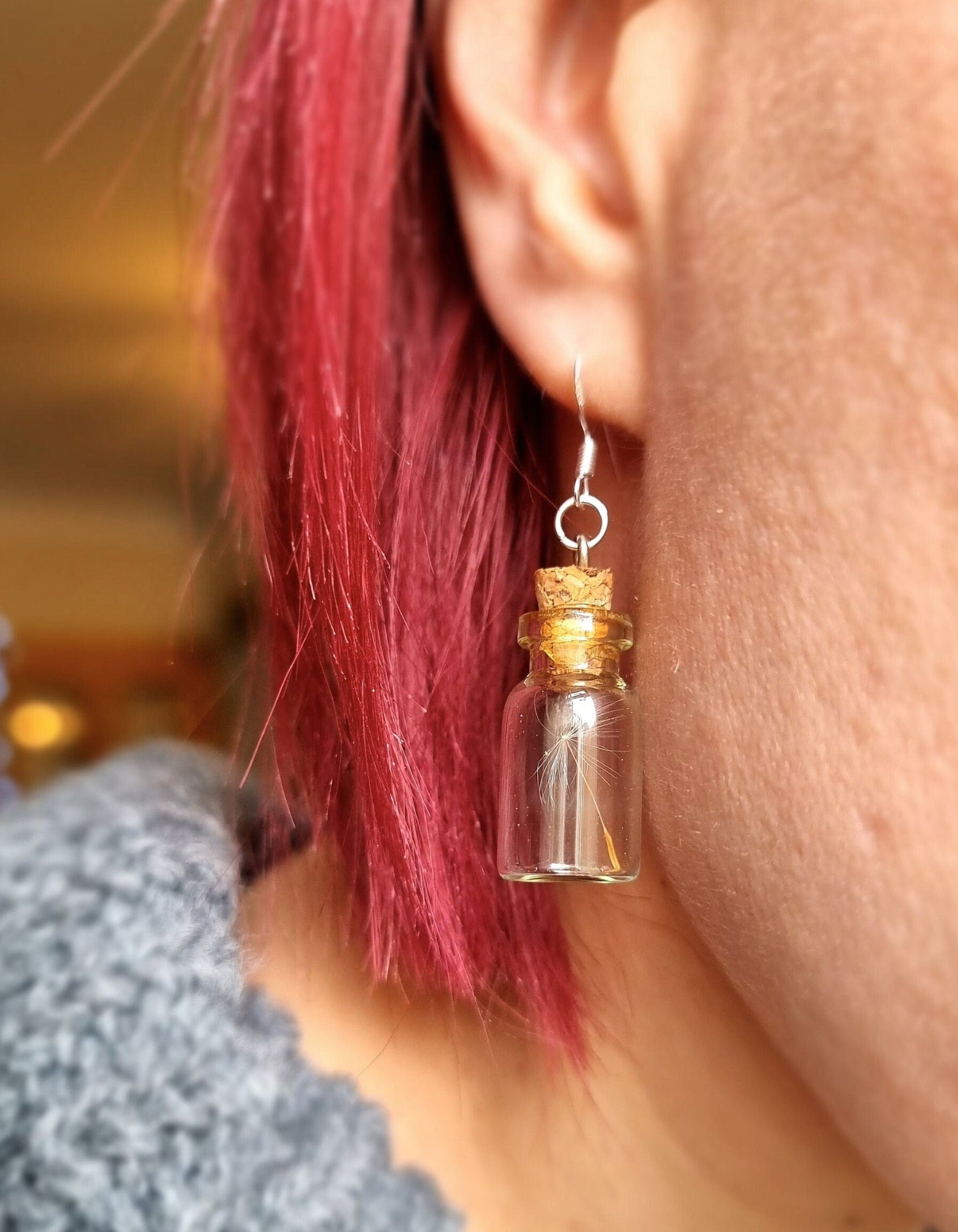 Jar earring containing dandelion seed hung from the ear lobe of a person with purple hair and a gret top