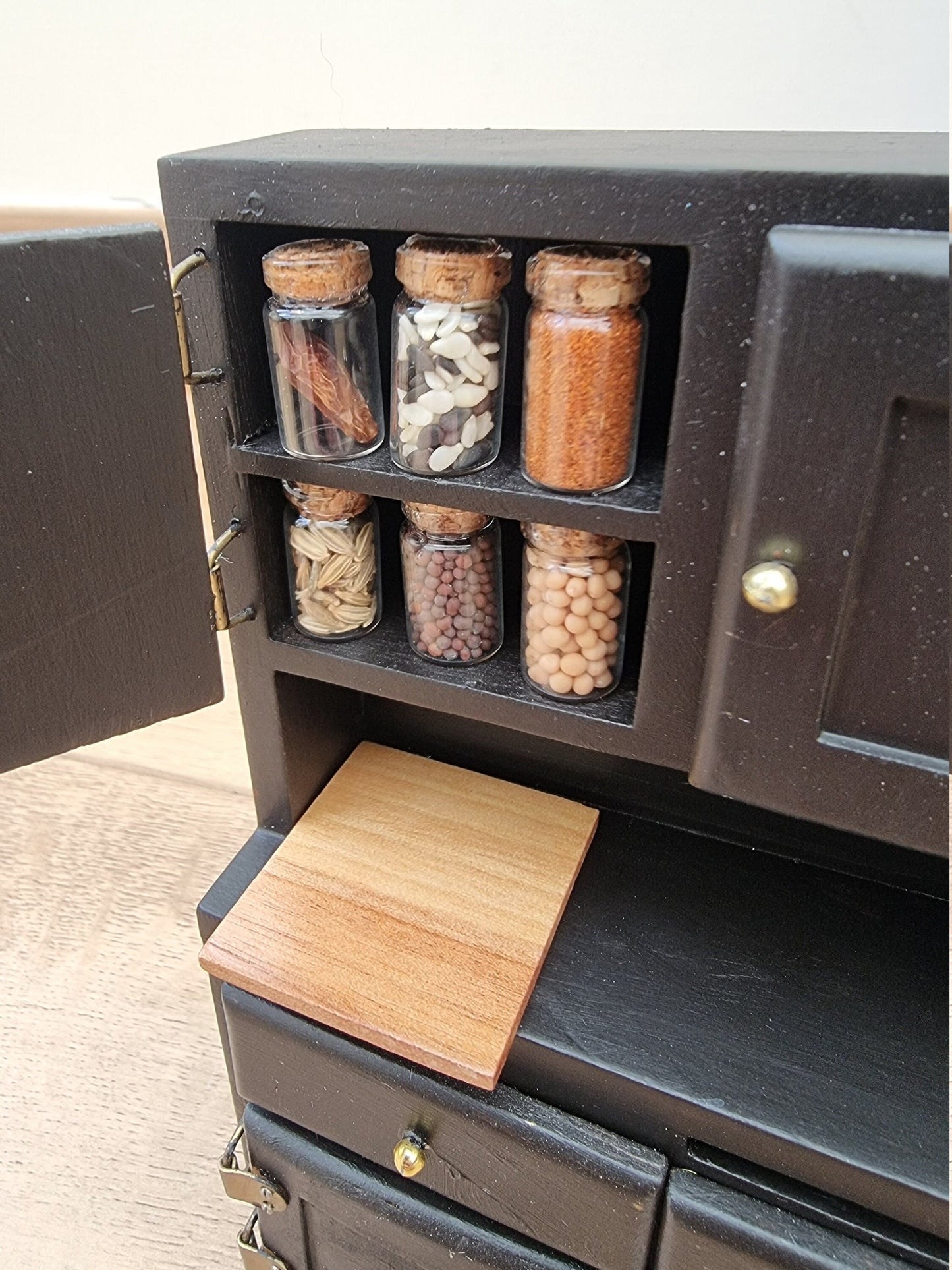 Set of 1:12 scale miniature apothecary jars with cork lids displayed in a black cupboard. Jars contain different herbs and spices