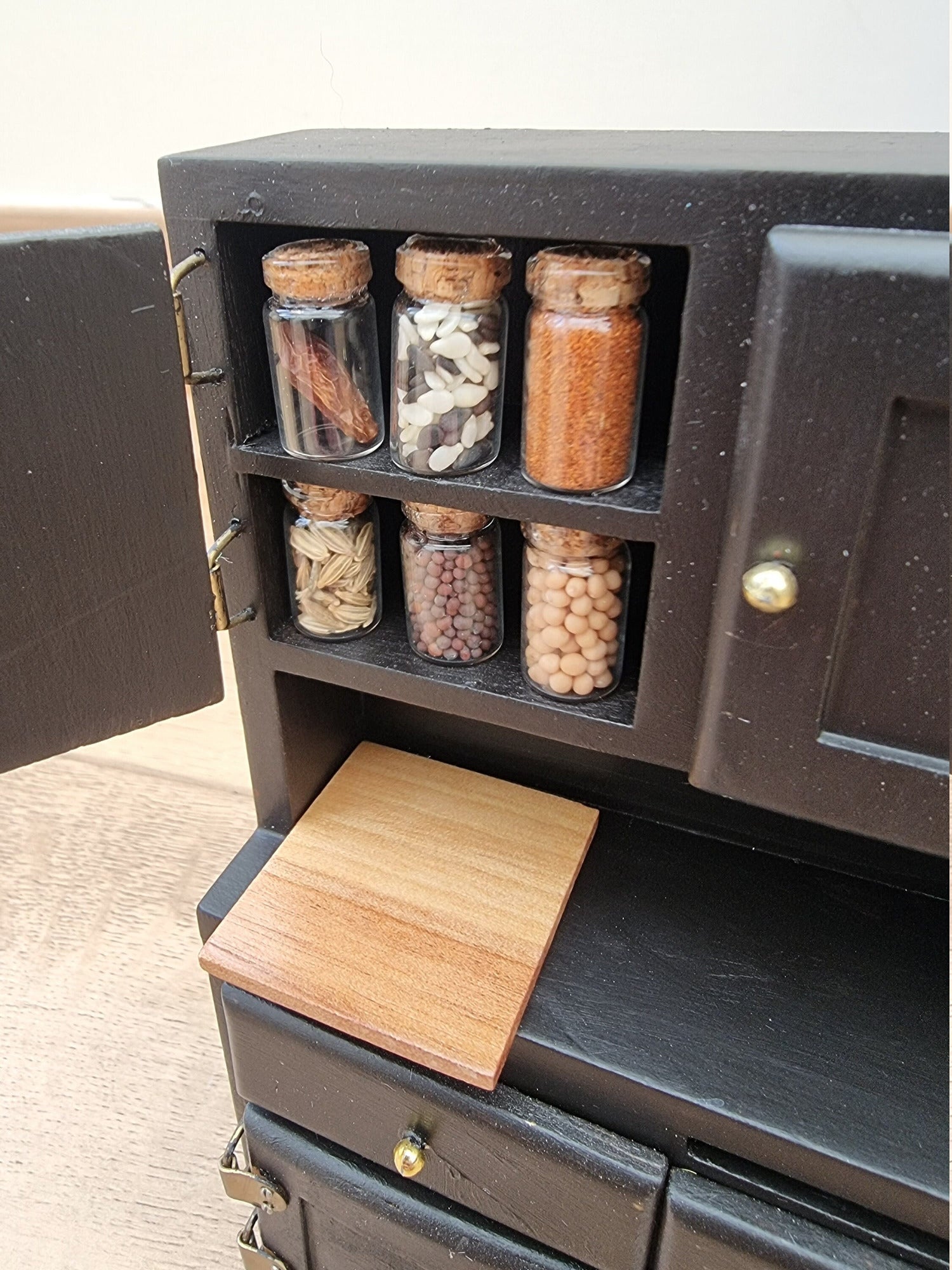 Set of 1:12 scale miniature apothecary jars with cork lids displayed in a black cupboard. Jars contain different herbs and spices