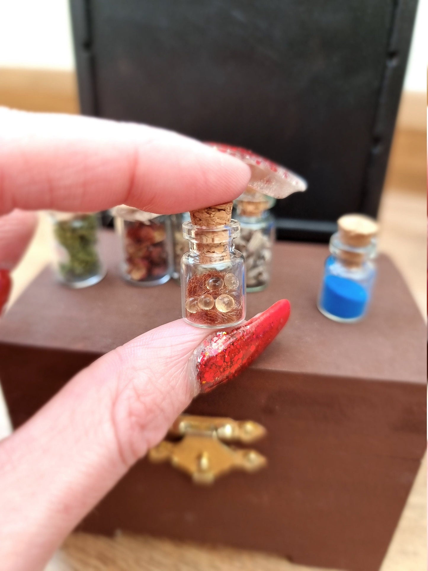 Small 1:12 scale apothecary jar containing clear balls and brown flockingwith a cork lid. Jar is held between a finger and thumb with other apothecary jars on a miniature chest in the background