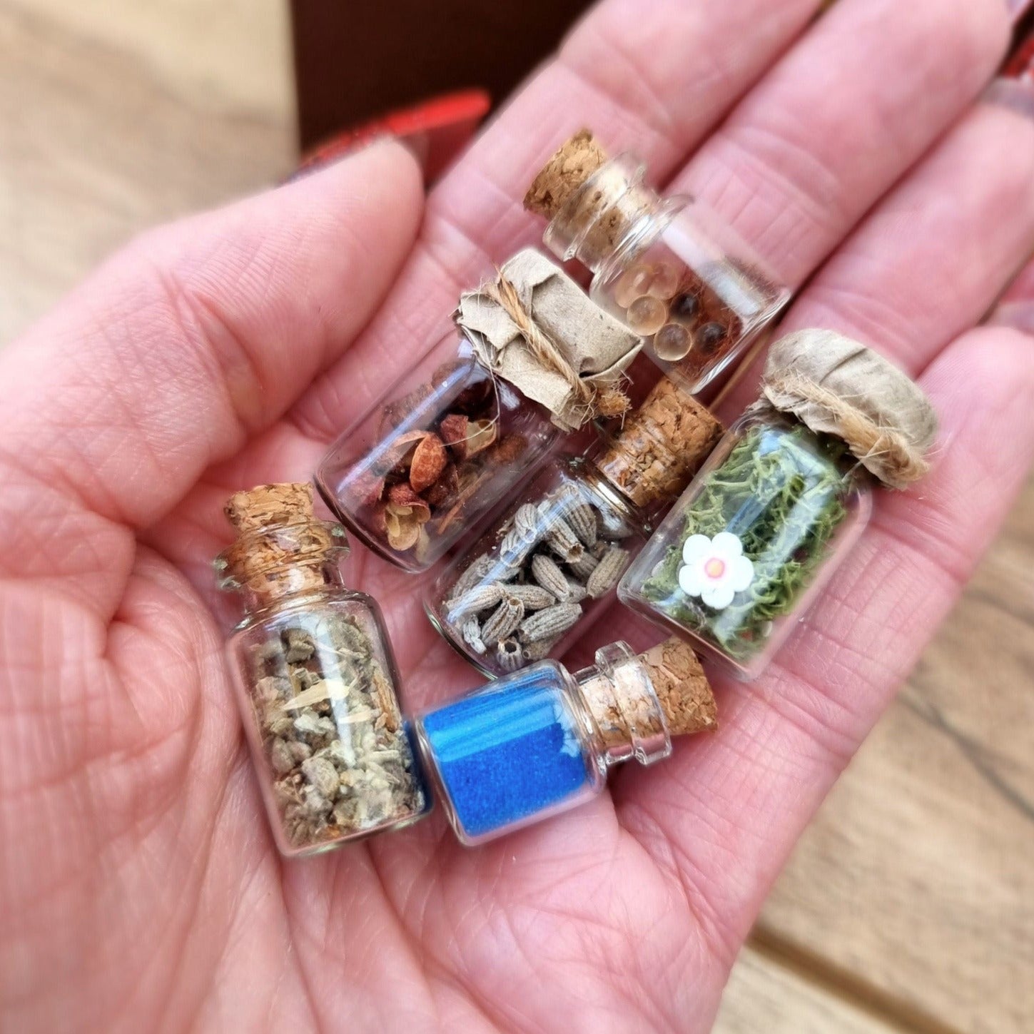 Set of 1:12 scale miniature apothecary jars with cork and paper lids held in an open palm. Jars contain blue powder, green herbs, red herbs, lavender herbs, green foliage with white flowers and clear balls brown flocking