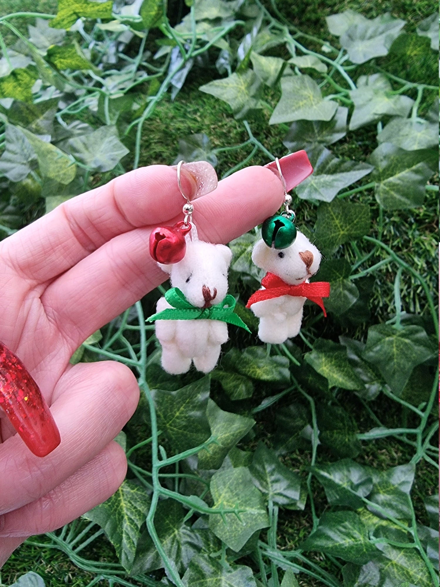 Two silver coloured hook style earrings hang from a hand. One hook has a small green bell and one has a small red bell. Each hook has a cream coloured fabric teddy bear with an alternate coloured ribbon bow tie to the bell