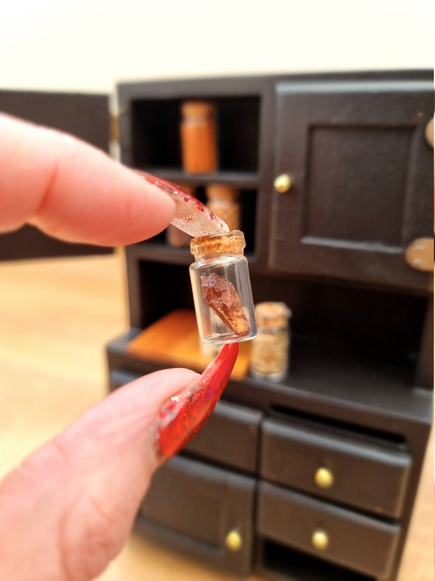 Single 1:12 scale apothecary jar with cork lid containing dried brown chilli. Jar is held between finger and thumb. Other jars are displayed in a black cupboard in the background and contain different herbs and spices