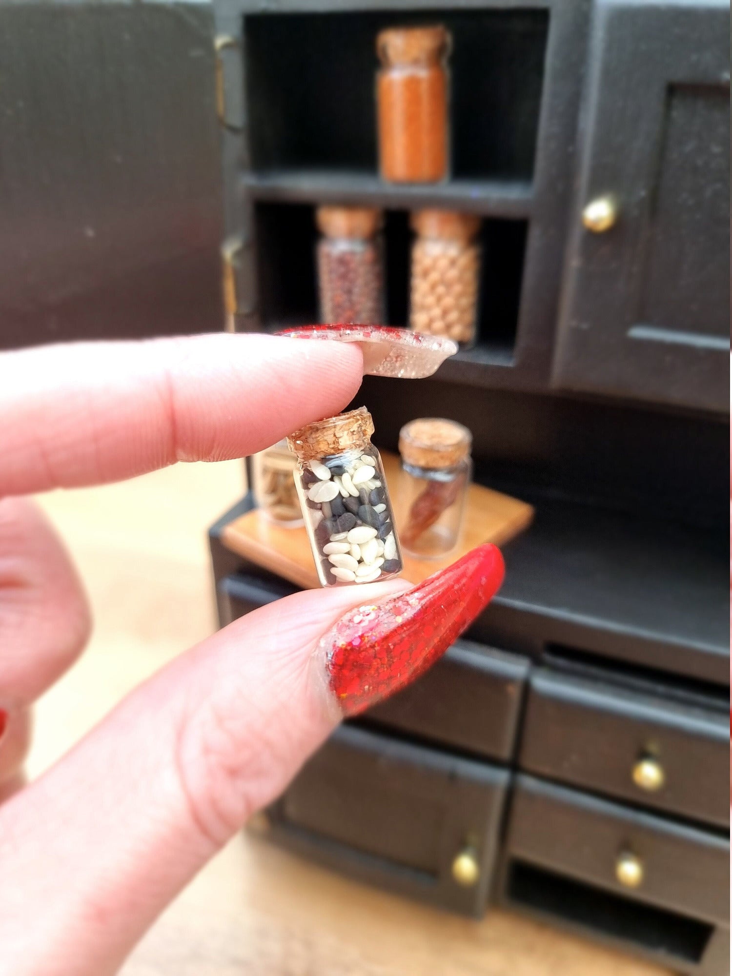 Single 1:12 scale apothecary jar with cork lid containing black and white seeds. Jar is held between finger and thumb. Other jars are displayed in a black cupboard in the background and contain different herbs and spices