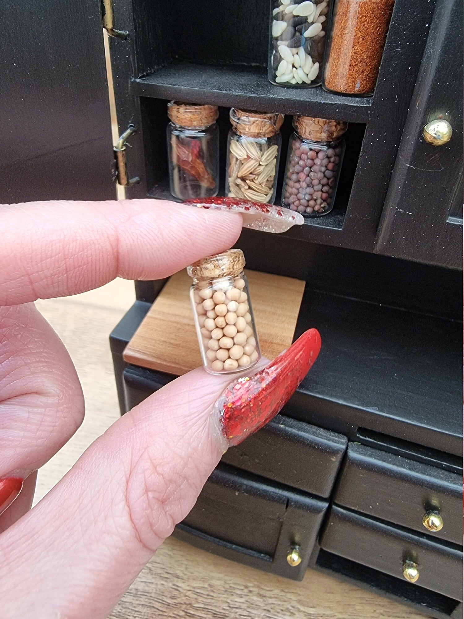 Single 1:12 scale apothecary jar with cork lid containing light yellow balls. Jar is held between finger and thumb. Other jars are displayed in a black cupboard in the background and contain different herbs and spices