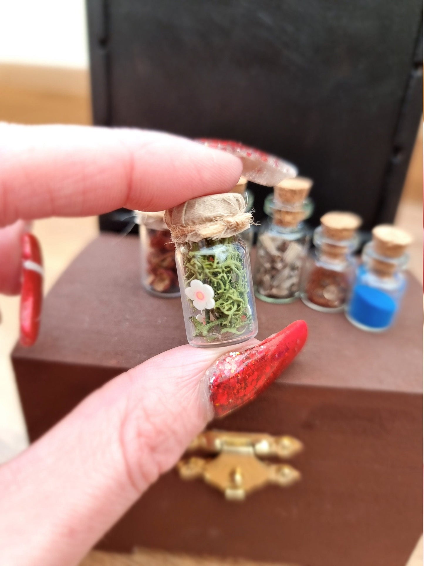 Miniature 1:12 scale apothecary jar with paper lid containing green foliage with white flowers. Jar is held between finger and thumb with other apothecary jars on a brown chest in the background