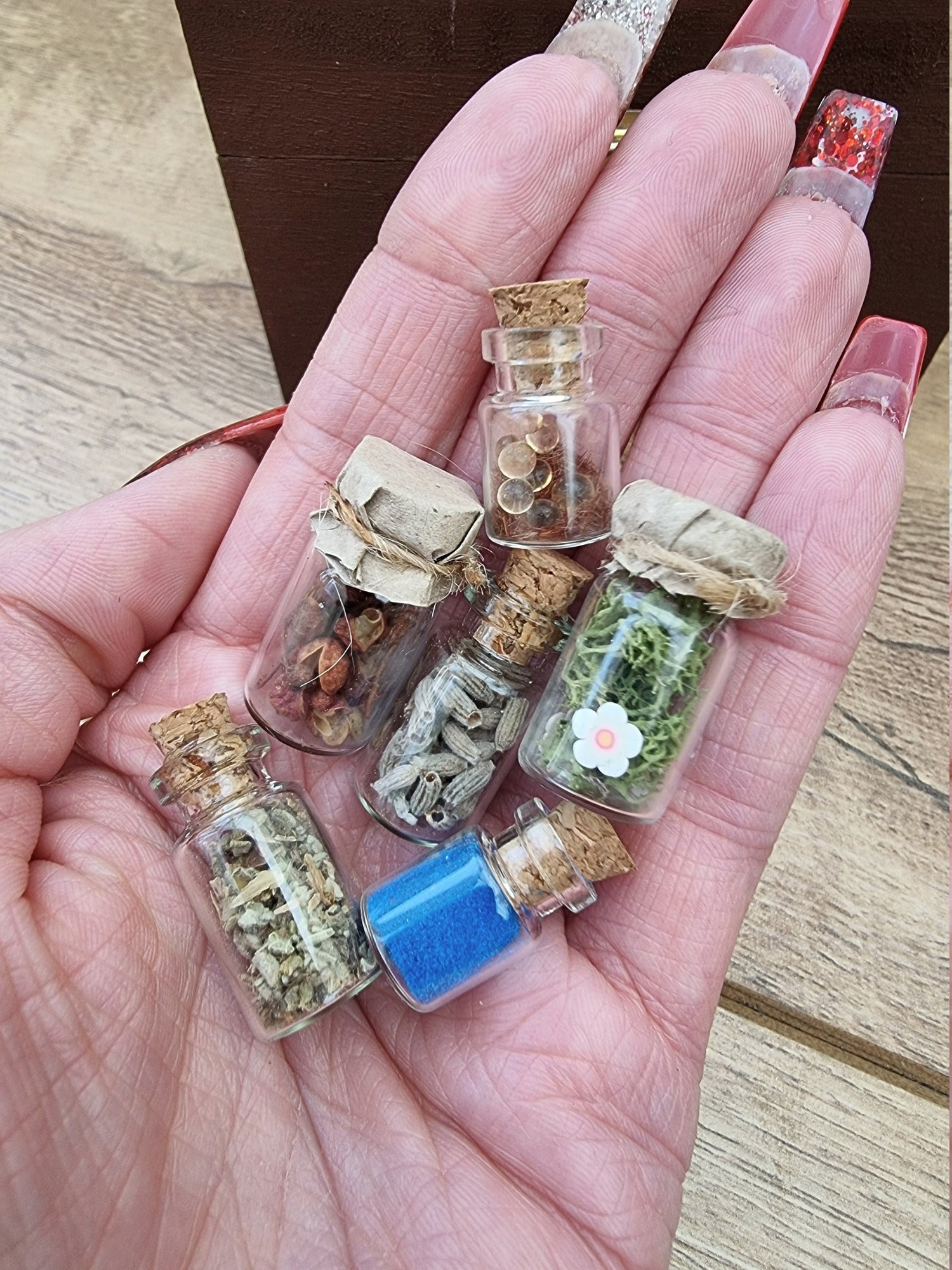 Set of 1:12 scale miniature apothecary jars with cork and paper lids held in an open palm. Jars contain blue powder, green herbs, red herbs, lavender herbs, green foliage with white flowers and clear balls brown flocking