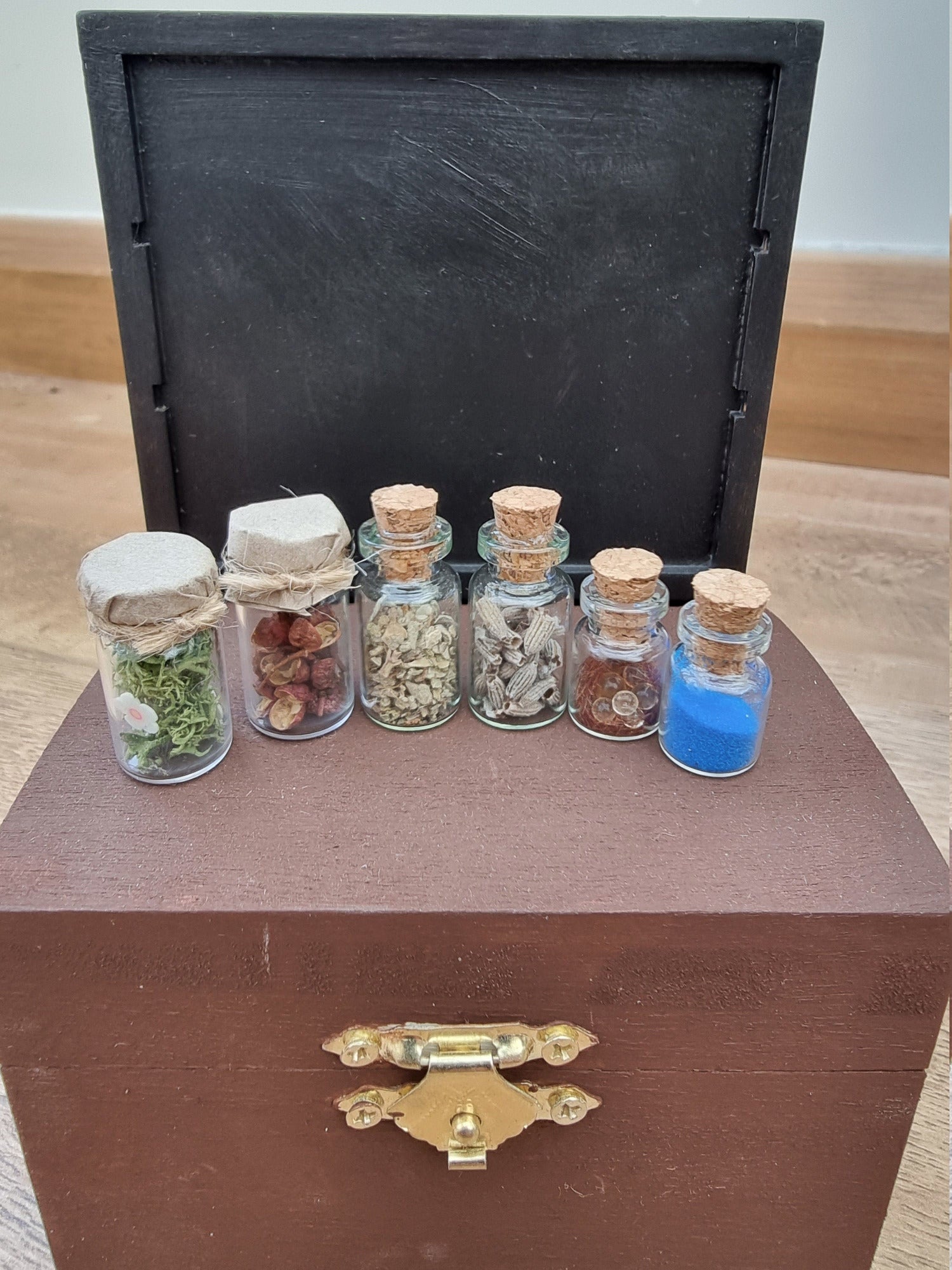 Set of 1:12 scale miniature apothecary jars with cork and paper lids on a brown chest. Jars contain blue powder, green herbs, red herbs, lavender herbs, green foliage with white flowers and clear balls brown flocking