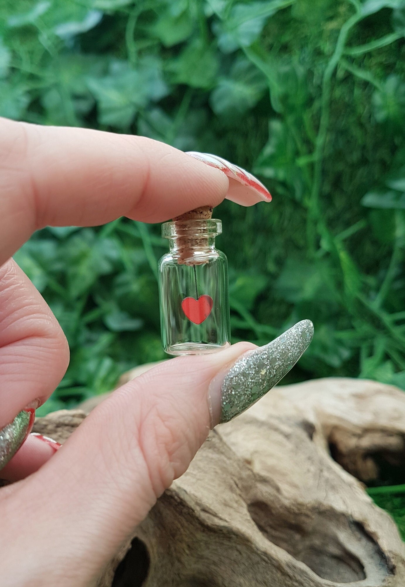 A miniature glass jar with a cork lid is held between fingers and thumb shown against a green ivy background. The tiny jar contains a red sequin heart hanging on black cotton from the cork inside the jar.