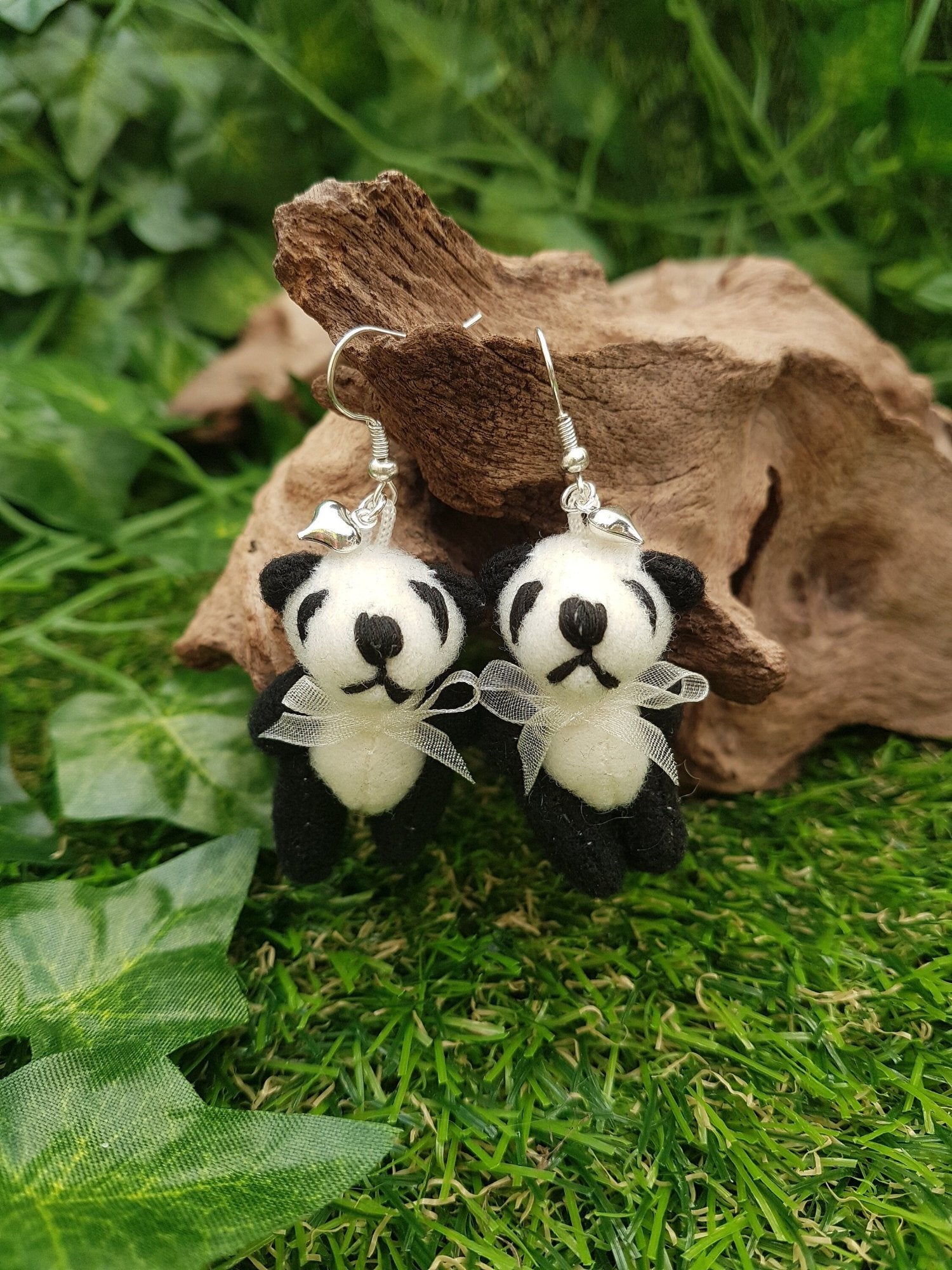 2 silver coloured hook style earrings displayed on driftwood, each with a small silver coloured fluffy heart charm and a larger black and cream coloured fabric panda teddy bear. The pandas wear a cream organza ribbon bow tie.