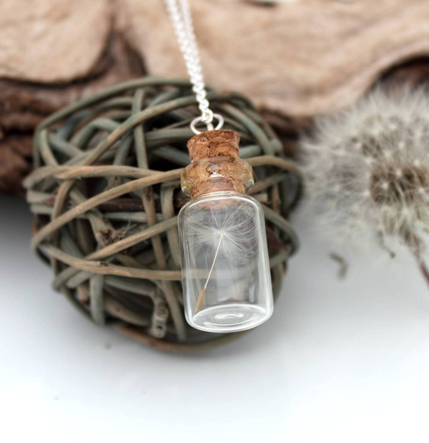 A glass vial necklace contains a single dandelion seed. The dandelion vial hangs on a fine sterling silver chain and is displayed on a grey wicker ball