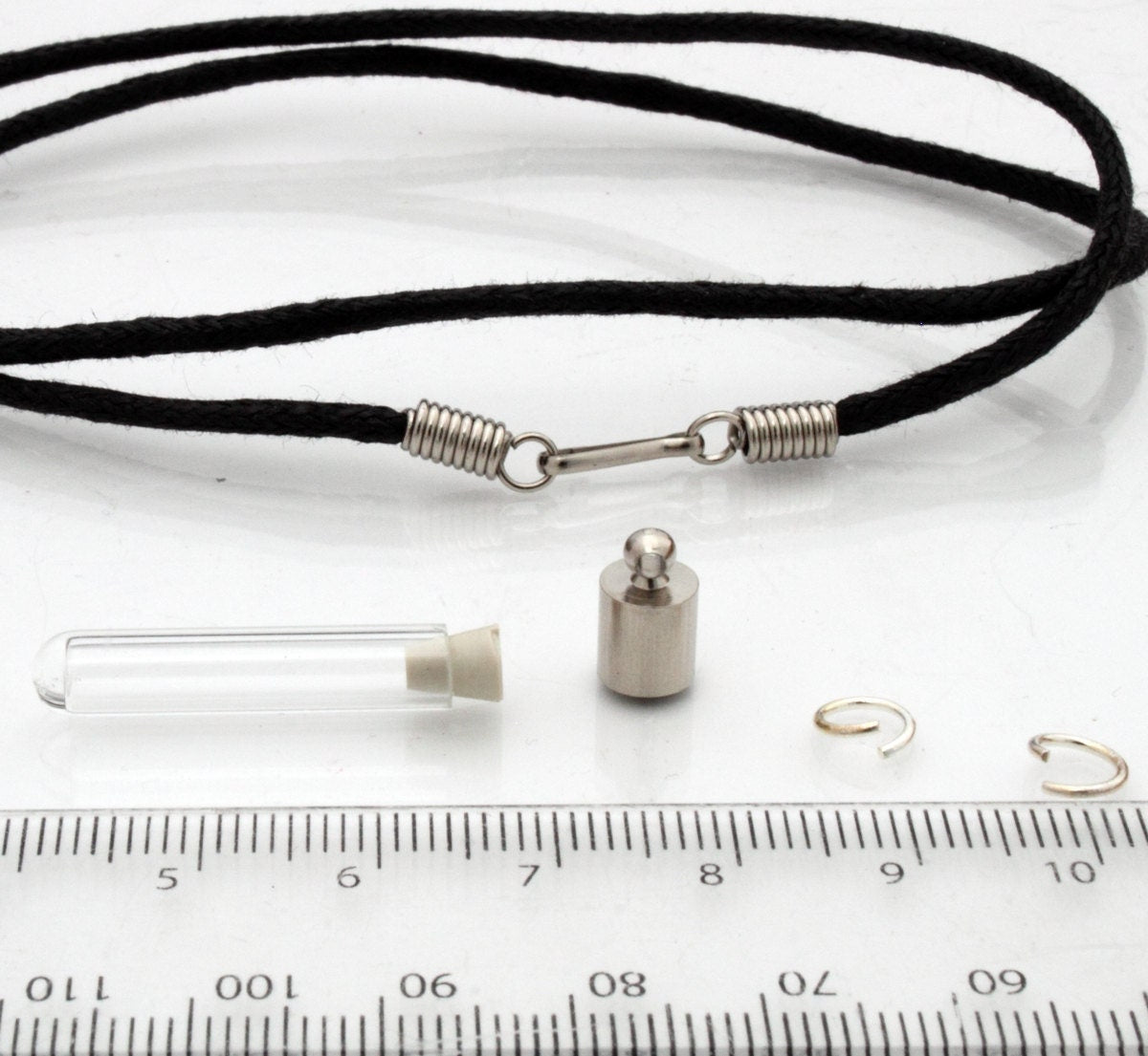 A miniature glass test tube shaped vial with silver coloured cap is shown on a white marble slab with a ruler for scale. 2 jump rings, a rubber bung and a black pendant cord seen in the background.