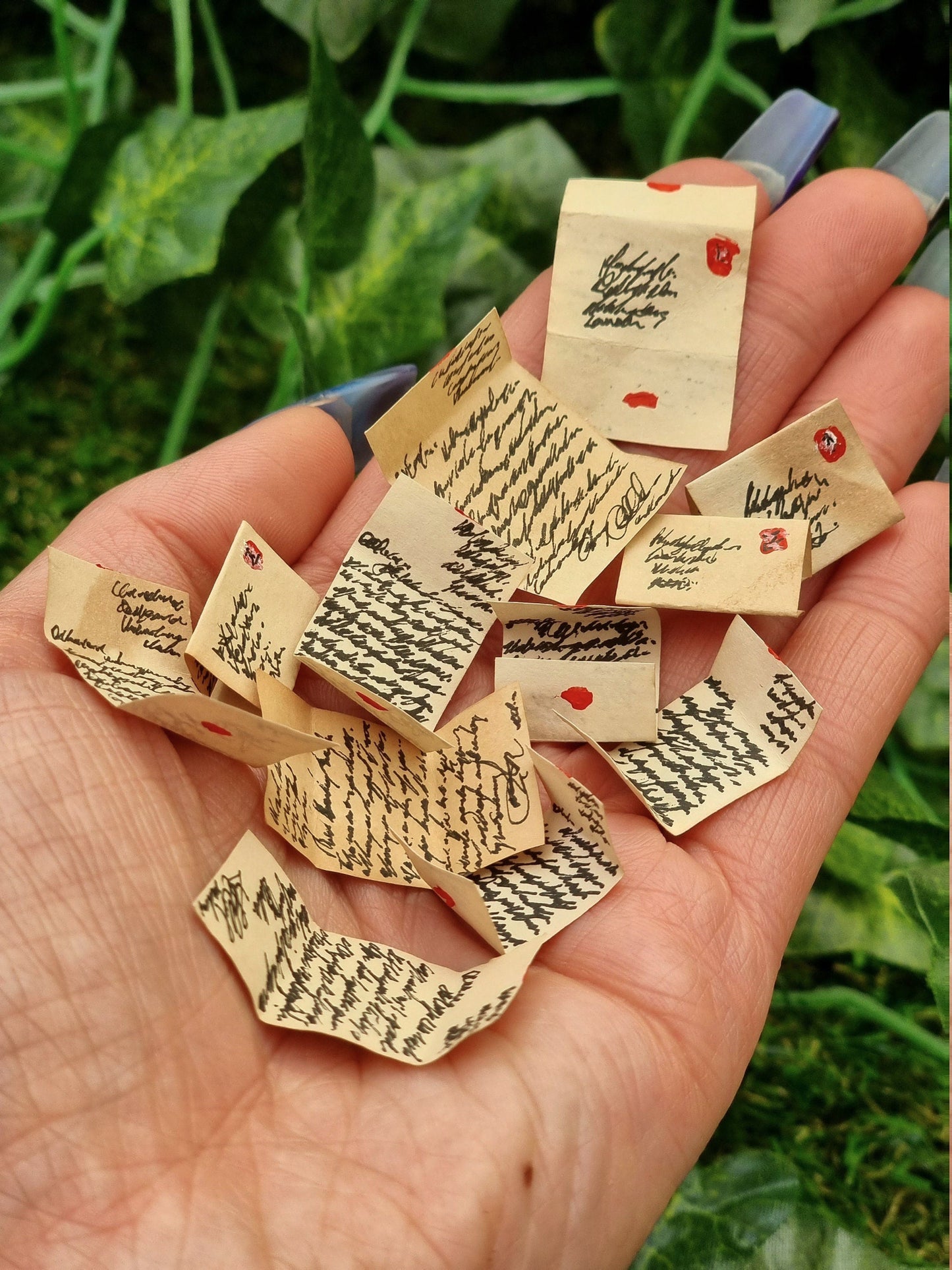 Lots of aged 1:12 scale miniature letters are displayed in the open palm of a hand. The letters are brown, showing black text. The letters are opened, so have folds, stamps, addresses and seals. Green ivy is seen in the background