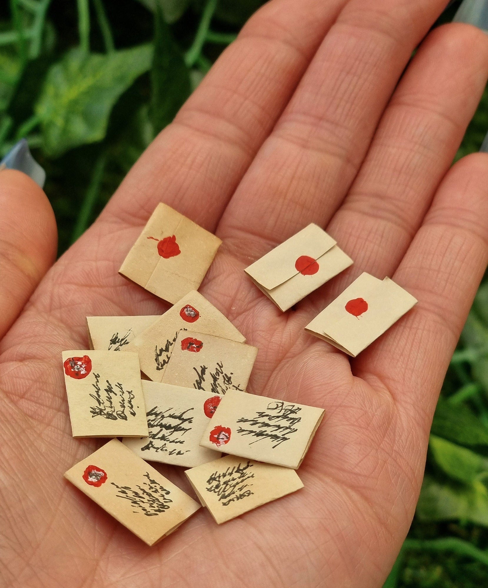 Lots of aged 1:12 scale miniature letters are displayed in the open palm of a hand. The letters are sealed, showing only a seal on the fold, stamp and address. Green ivy is seen in the background