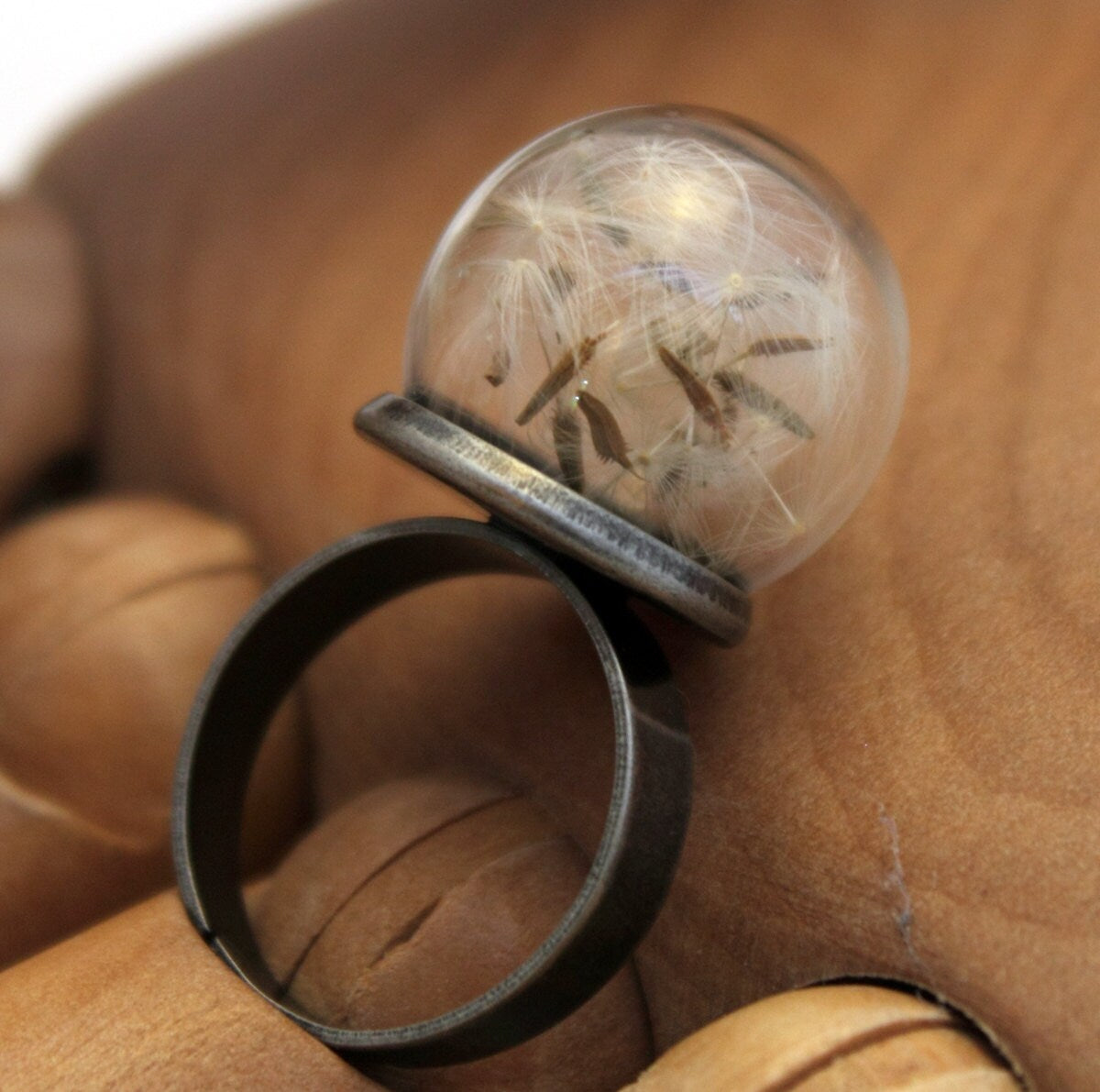 A glass globe ring containing delicate dandelion seeds is displayed on a wooden articulated hand. The ring is a gunmetal colour