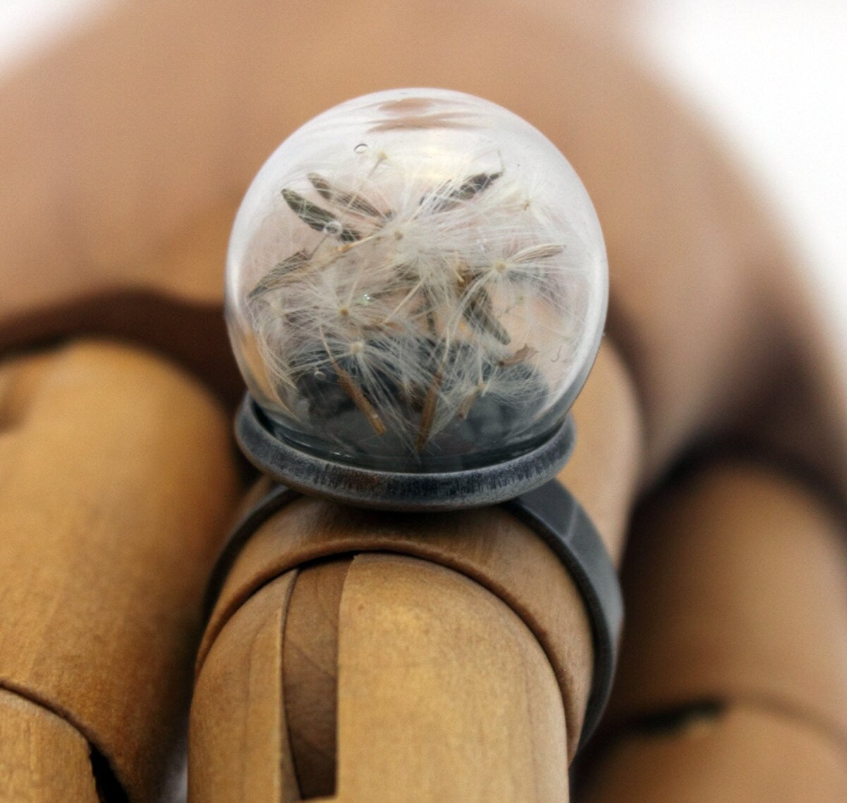 A glass globe ring containing delicate dandelion seeds is displayed on a wooden articulated hand. The ring is a gunmetal colour