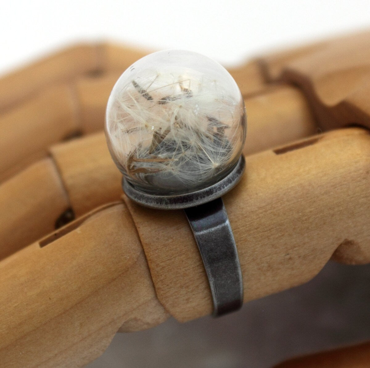 A glass globe ring containing delicate dandelion seeds is displayed on a wooden articulated hand. The ring is a gunmetal colour