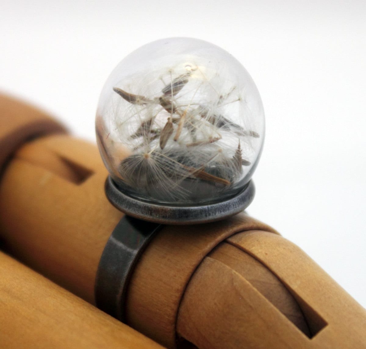 A glass globe ring containing delicate dandelion seeds is displayed on a wooden articulated hand. The ring is a gunmetal colour