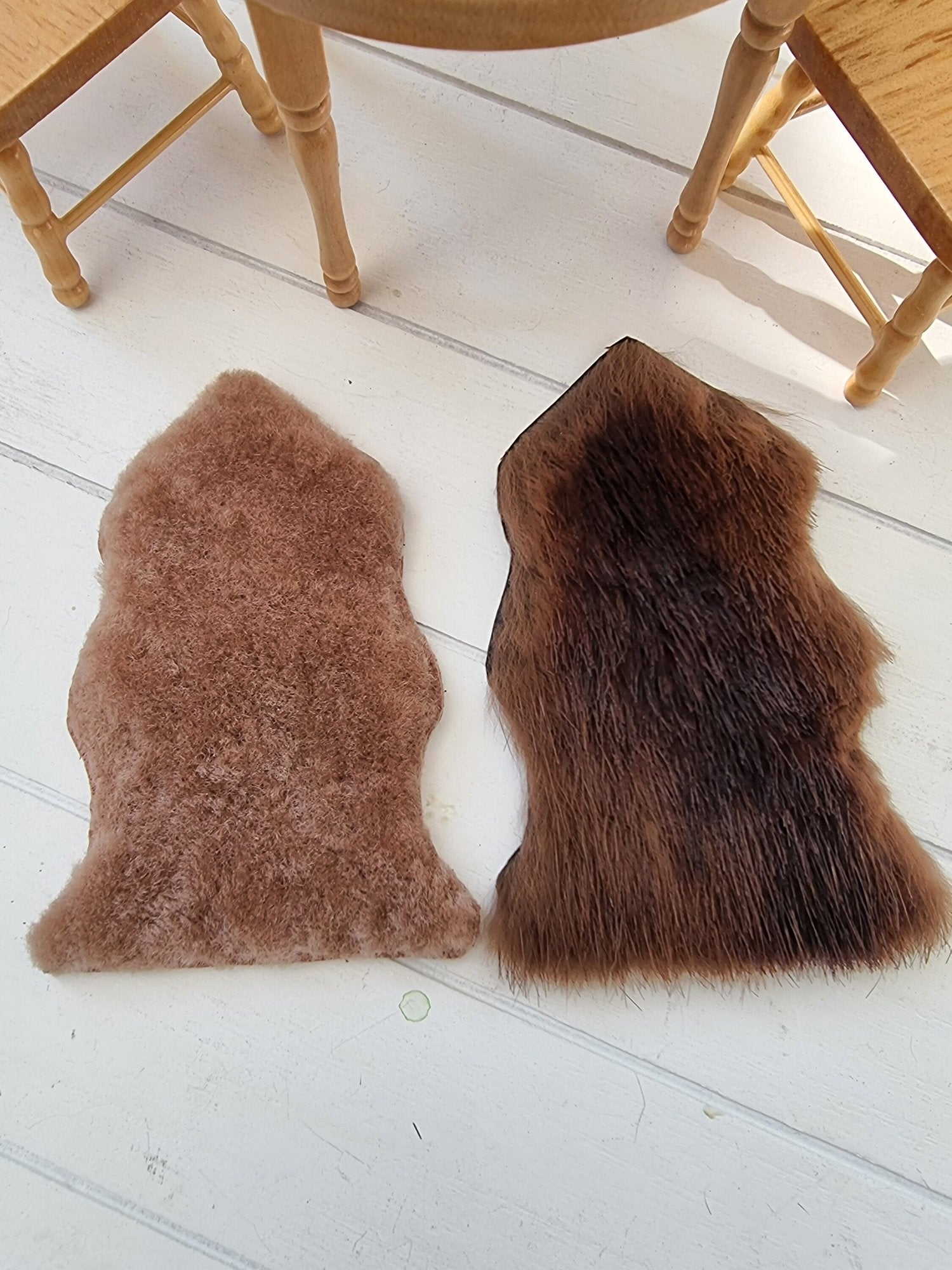 Two 1:12 scale animal skin genuine fur rugs lie on a white wooden floor with miniature table and chairs in the background. One is dark brown and the other is light brown.