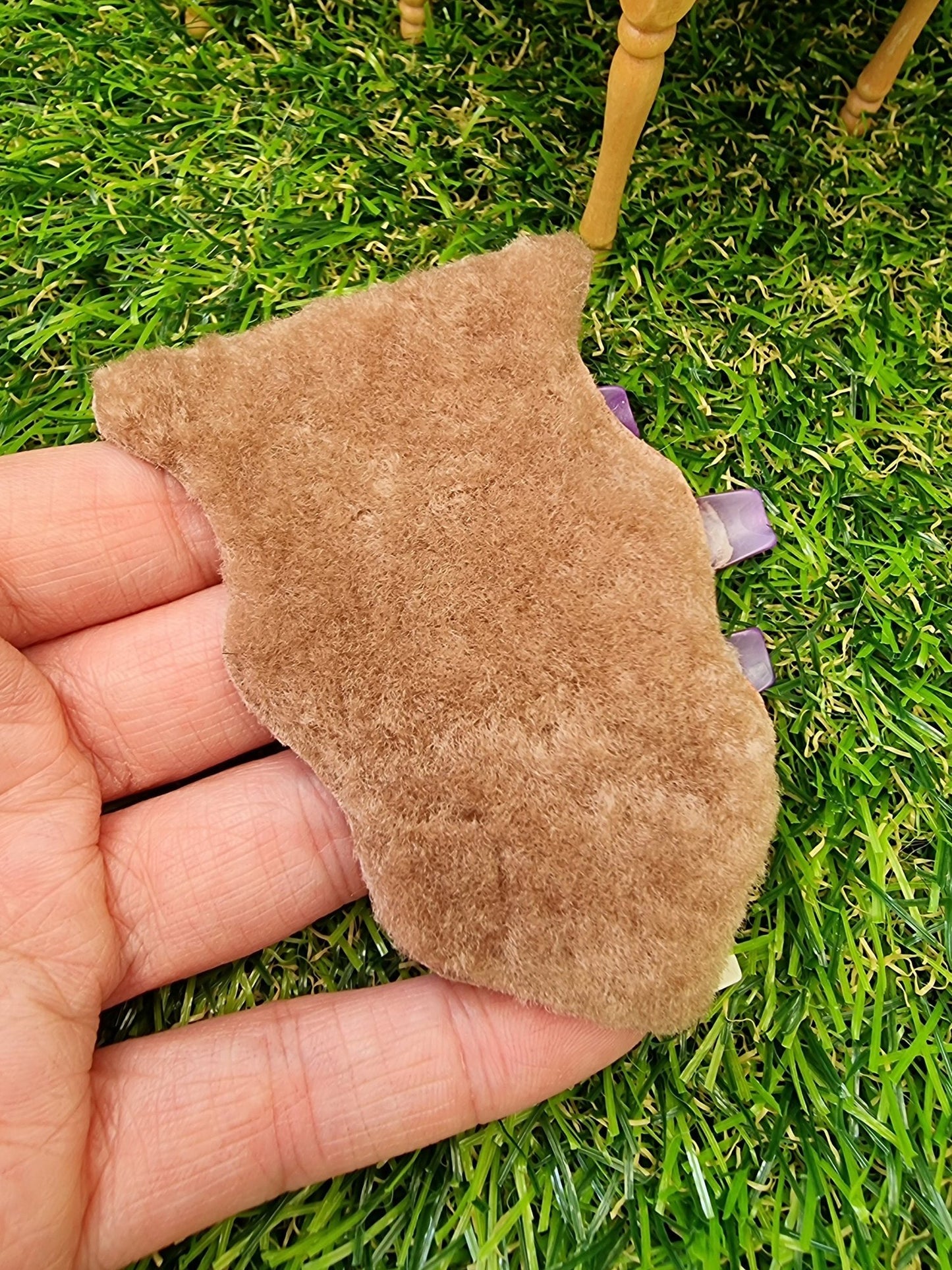 Hand holds a 1:12 scale light brown animal skin genuine fur rug on a green grass floor in the background. The rug is light brown with a short pile