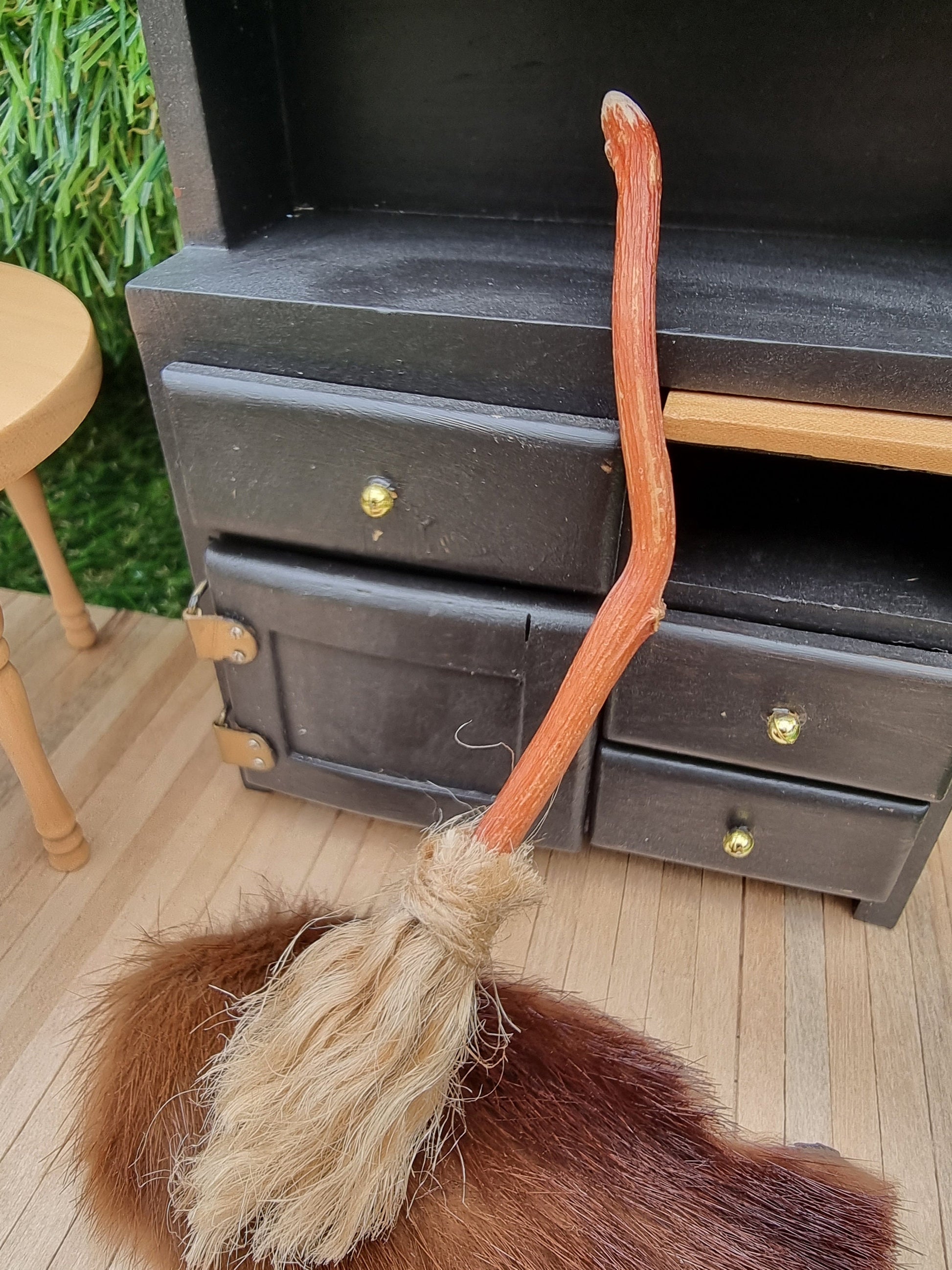 A small 1:12 broom leans against a black dresser and rug in the background. The broom has brown bristles and a brown bent handle for flying