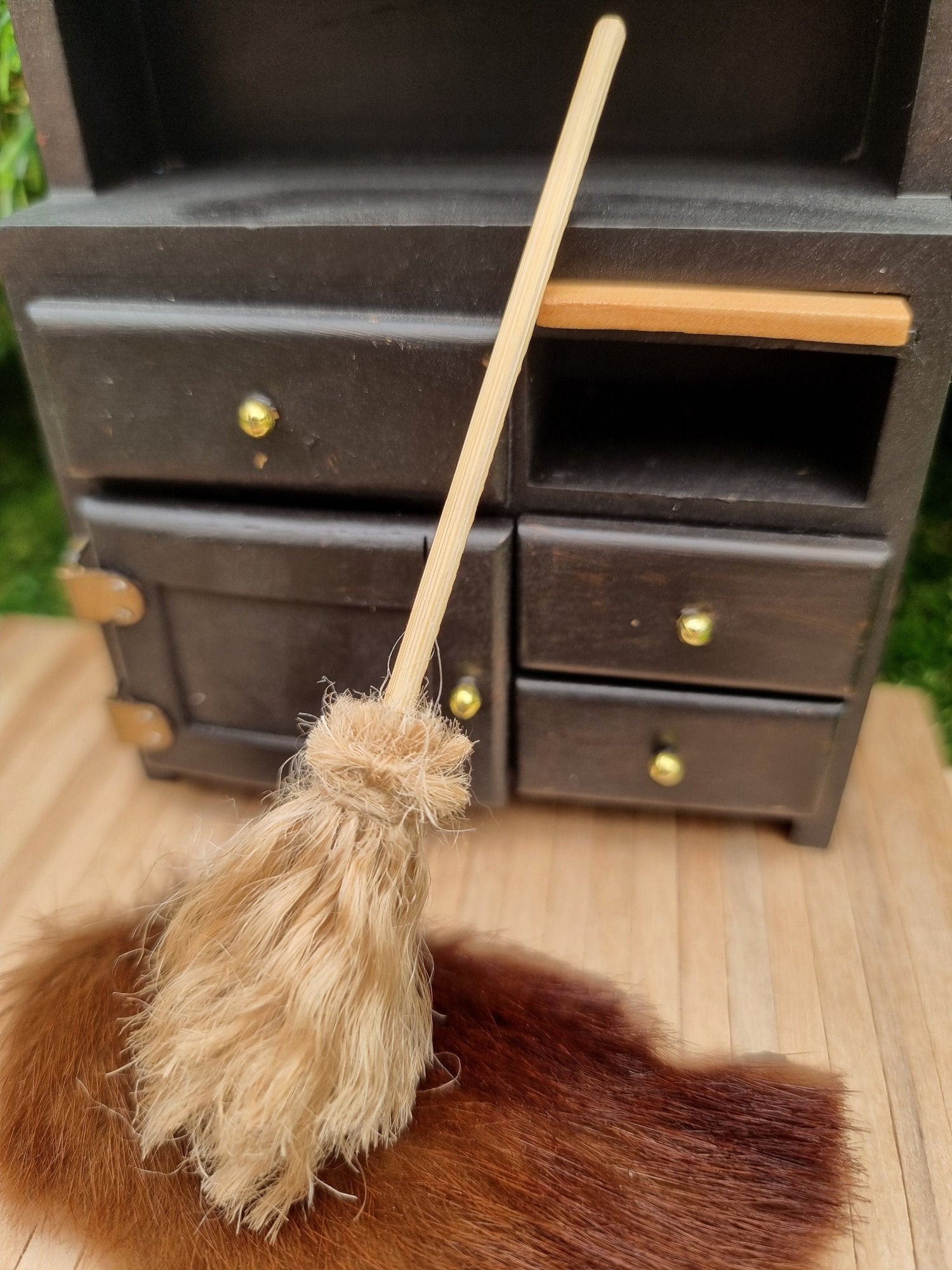 A small 1:12 broom leans against a black dresser and rug in the background. The broom has brown bristles and a long straight handle for sweeping