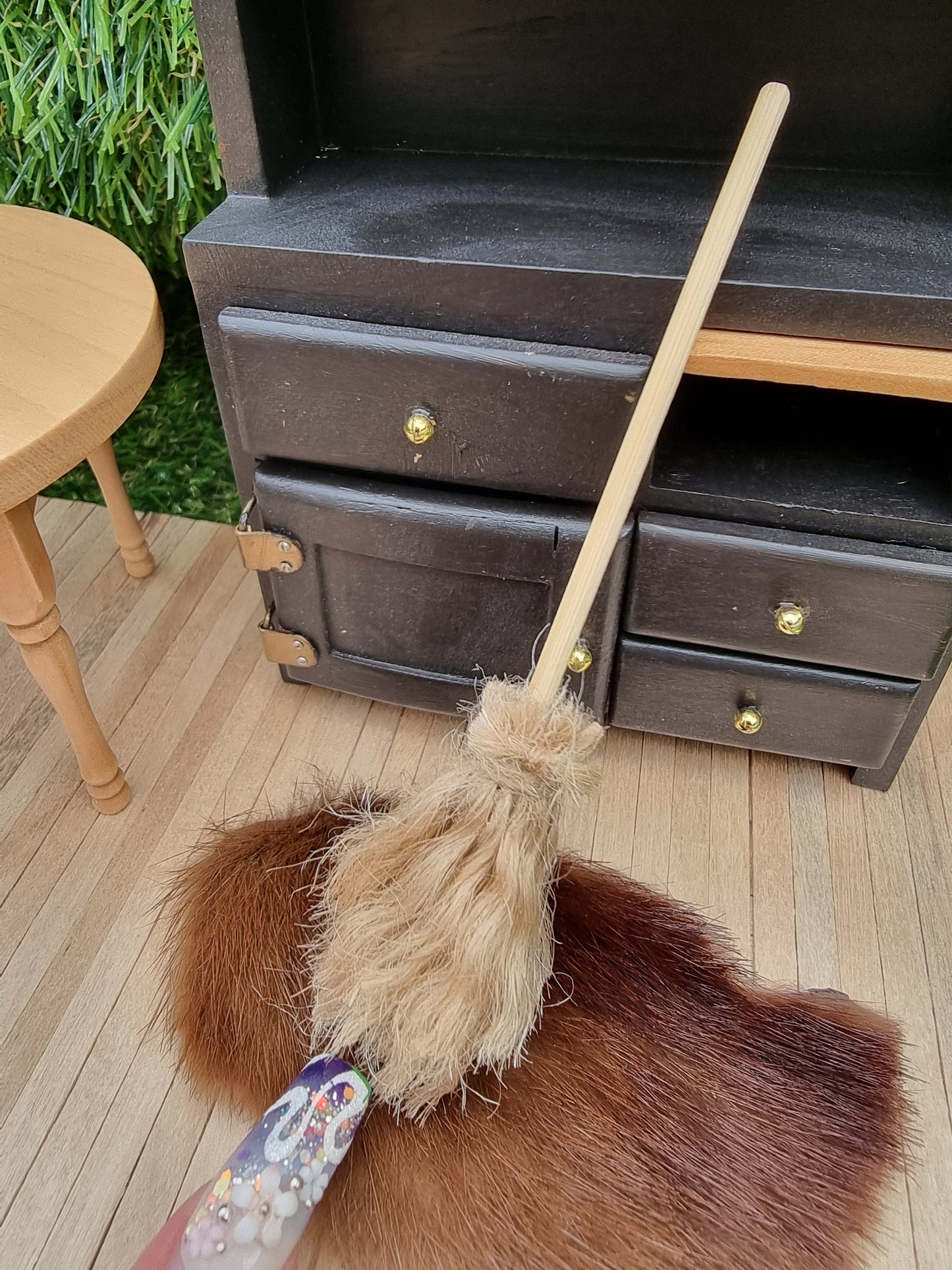 A finger points at a small 1:12 broom leaning against a black dresser and rug in the background. The broom has brown bristles and a long straight handle for sweeping