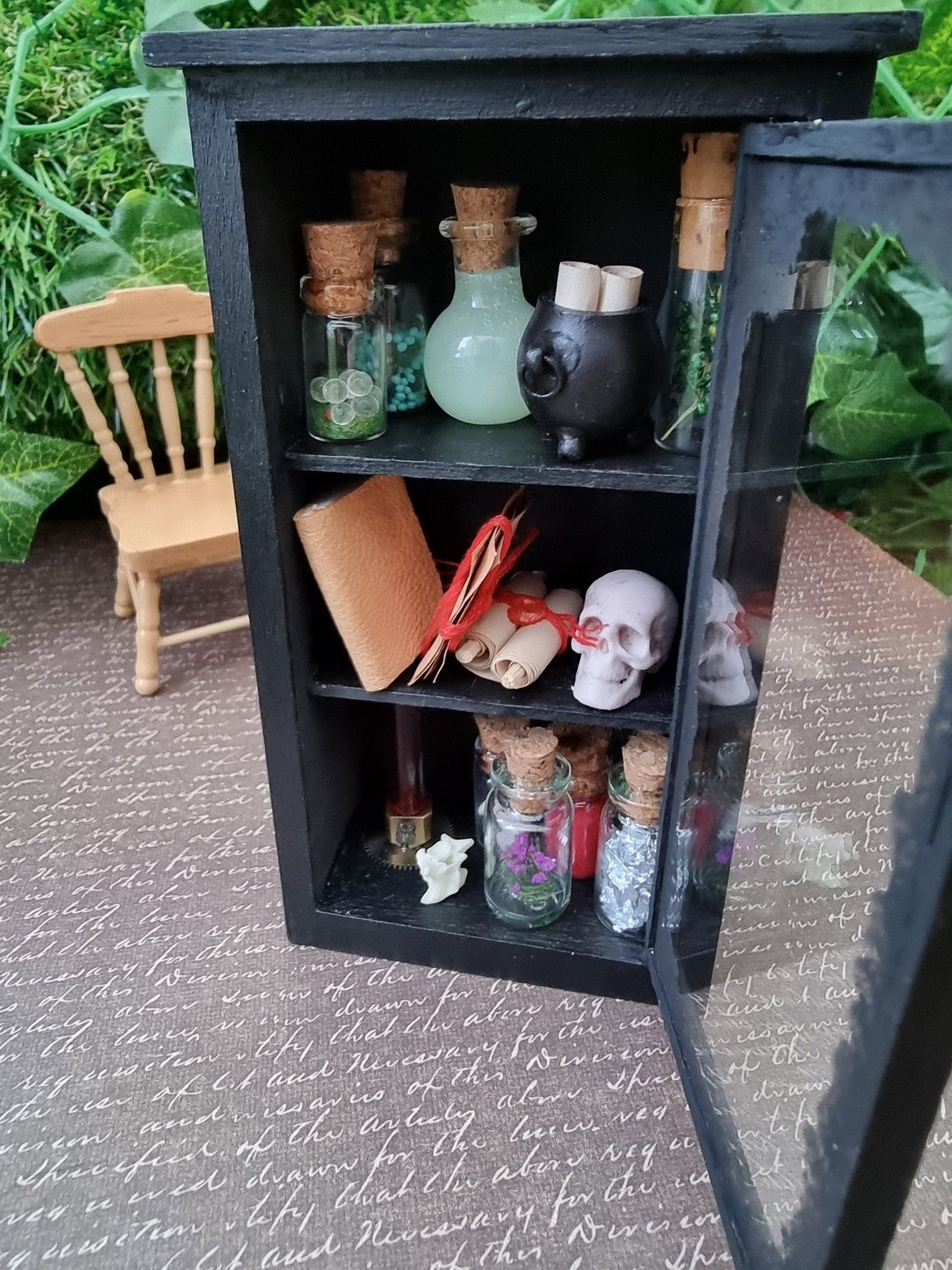 Open miniature 1:12 scale glass-fronted black apothecary cabinet stands against an ivy background. The cabinet has 3 shelves. The top shelf has apothecary bottles and a cauldron. Middle shelf has a book, skull and scroll. Bottom shelf has apothecary bottles