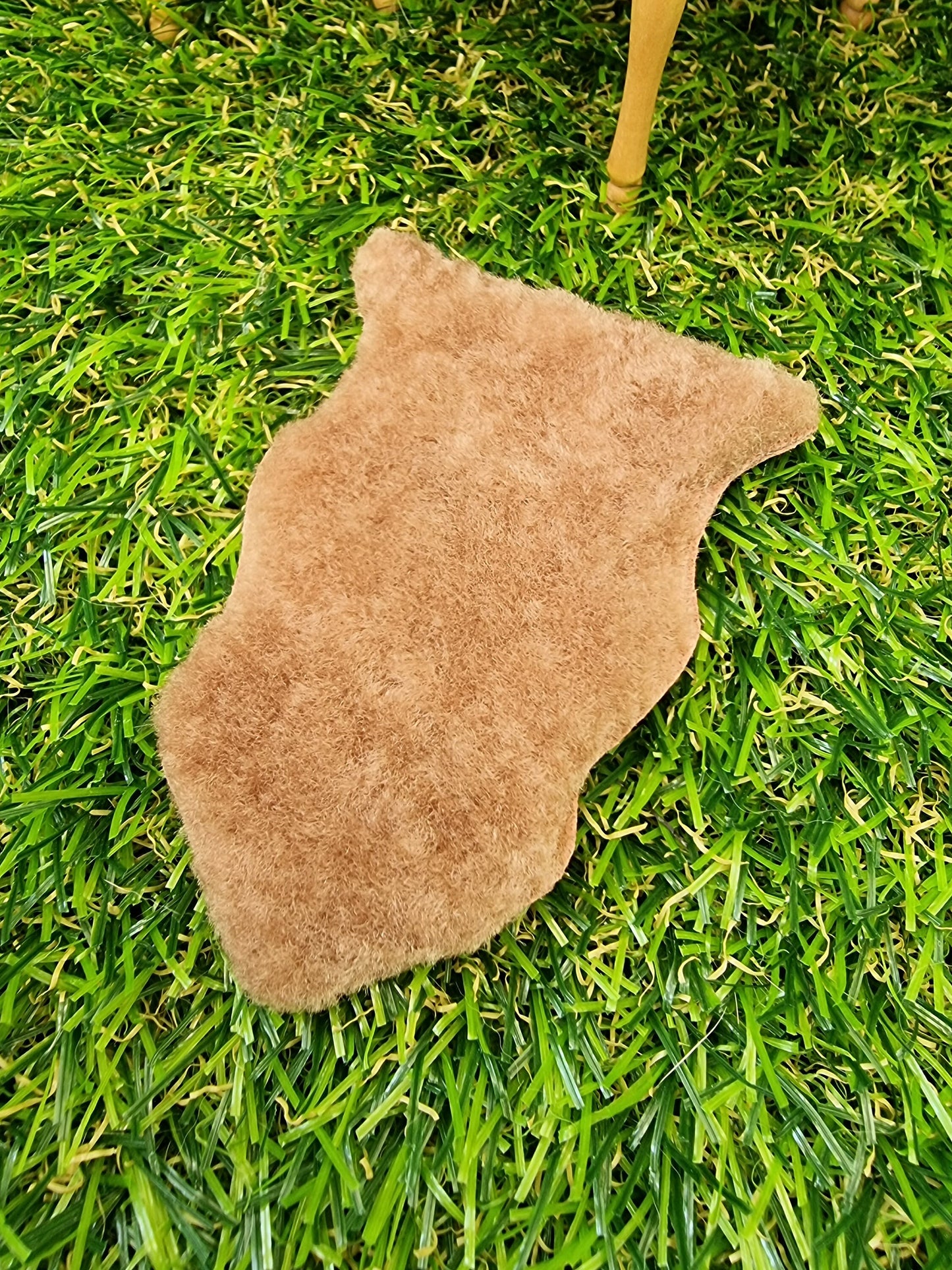 1:12 scale light brown animal skin genuine fur rug on a green grass floor with miniature table in the background. The rug is light brown with a short pile