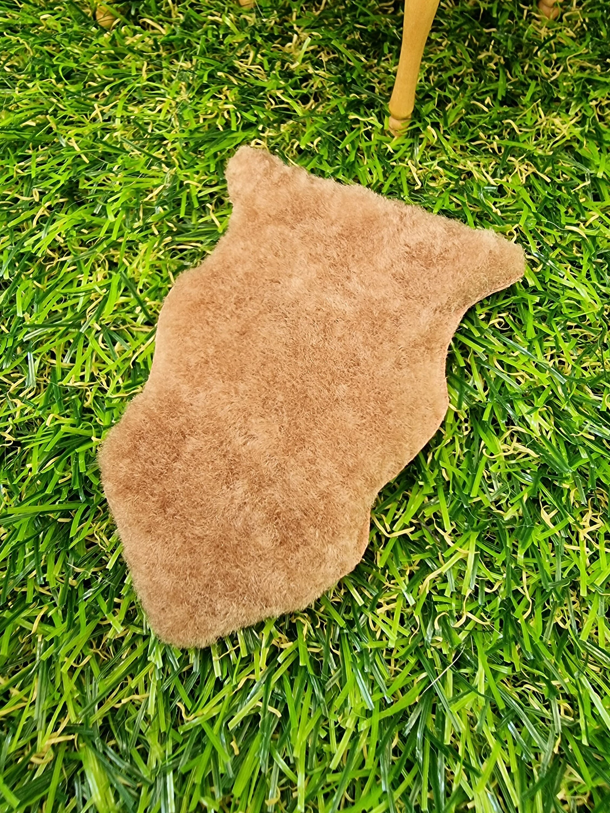 1:12 scale light brown animal skin genuine fur rug on a green grass floor with miniature table in the background. The rug is light brown with a short pile