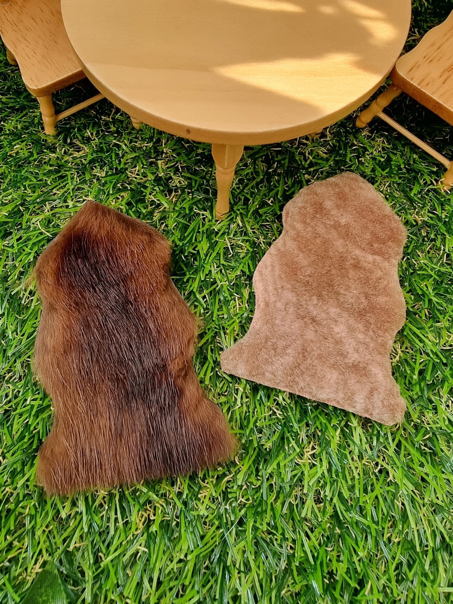 Two 1:12 scale animal skin genuine fur rugs lie on a green grass floor with miniature table and chairs in the background. One is dark brown and the other is light brown.