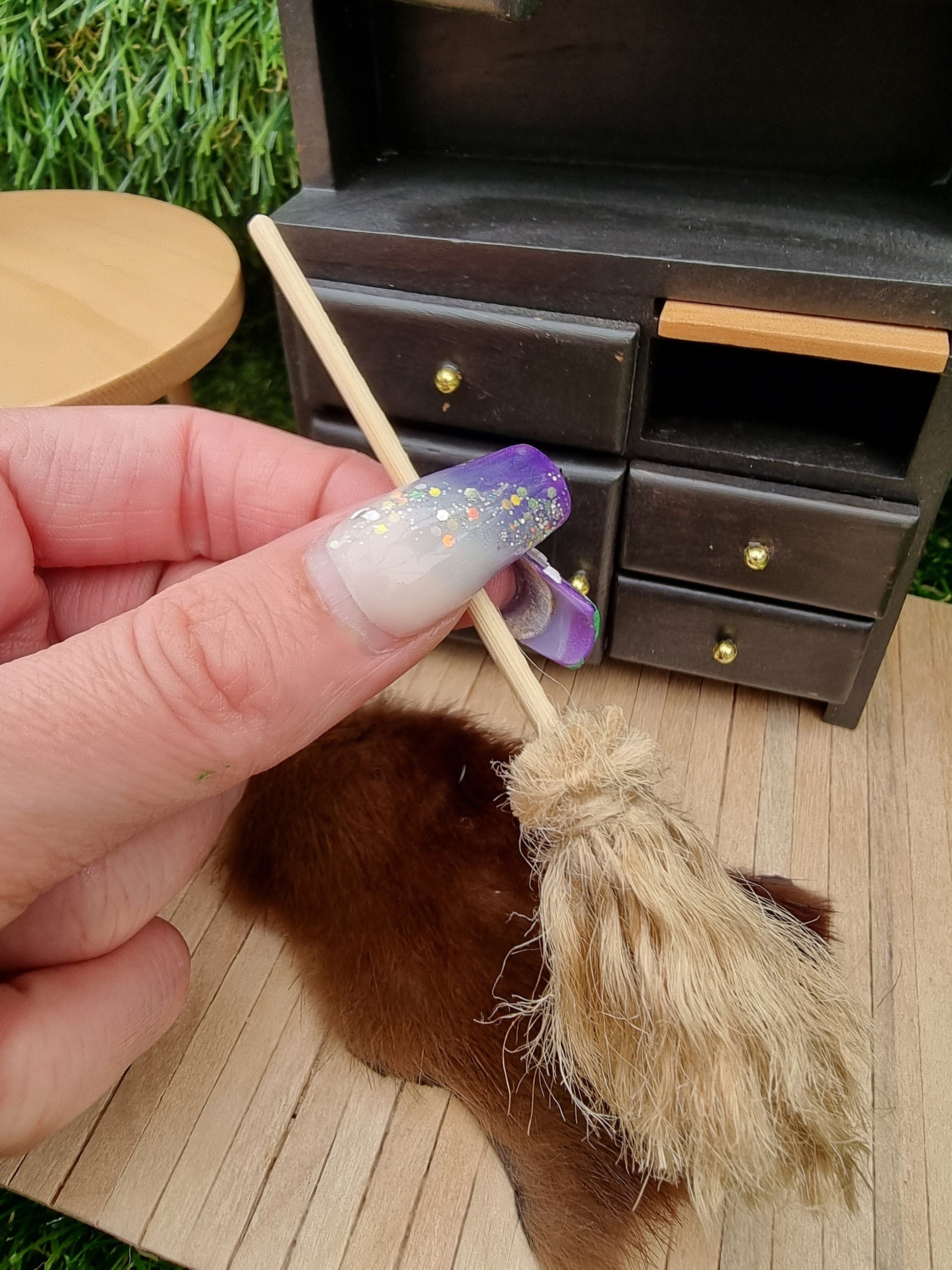 Fingers hold a small 1:12 broom with a black dresser and rug in the background. The broom has brown bristles and a long straight handle for sweeping