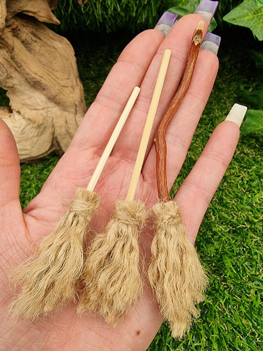 3 1:12 brooms sit in the palm of a hand. All brooms have brown bristles and handles. One broom has a shorter handle, one has a longer handle and one has a longer bent handle for flying