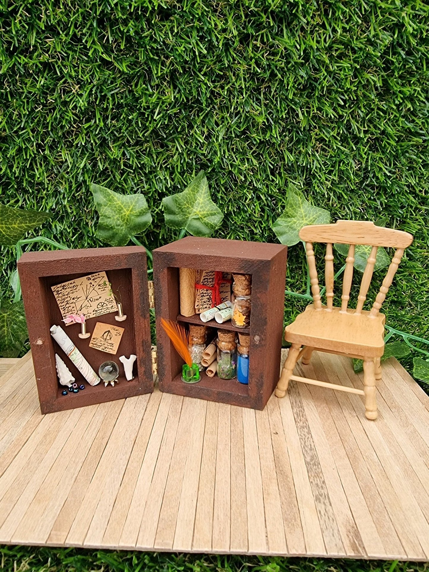 A 1:12 scale miniature brown apothecary cabinet is shown open on a marble slab with glass vials, parchment scrolls, miniature cauldron and leather bound book. The left cabinet door has aged notes pinned up with an inkwell and quill